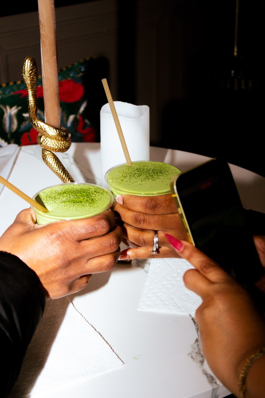 Two hands clinking green matcha lattes with bamboo straws on a white table.