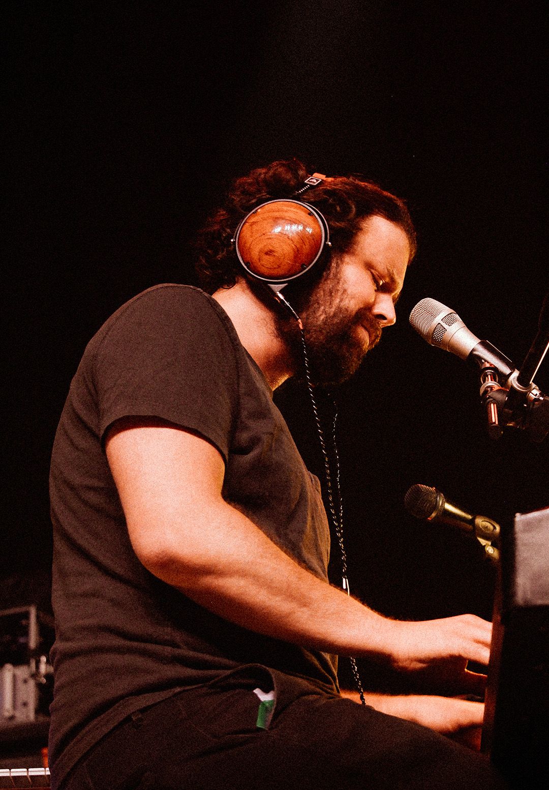 Bearded male musician wearing brown headphones plays piano at a microphone in a dark studio setting.
