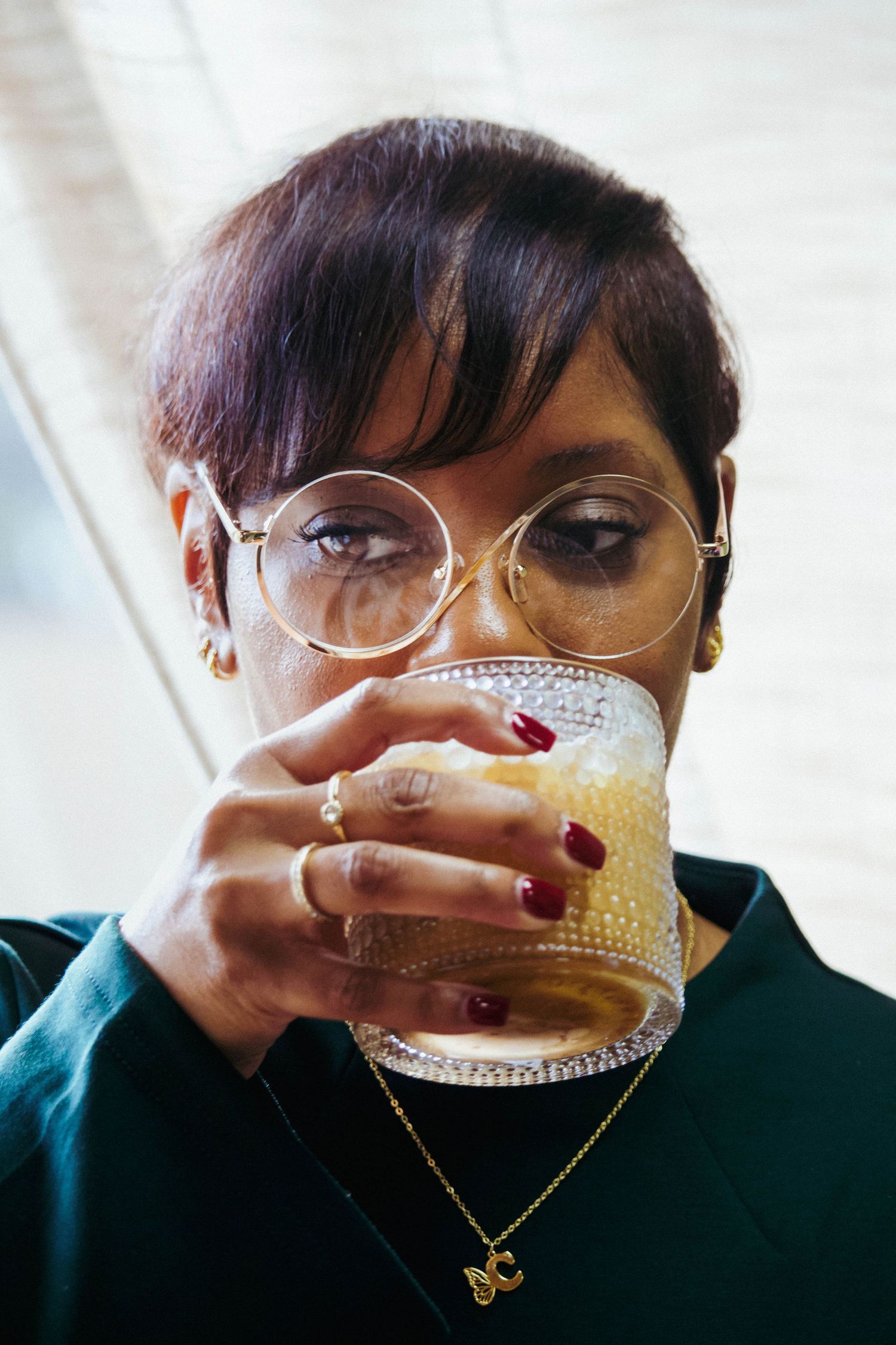 Woman with short hair and round glasses sipping a craft cocktail with red manicured nails and gold jewelry.