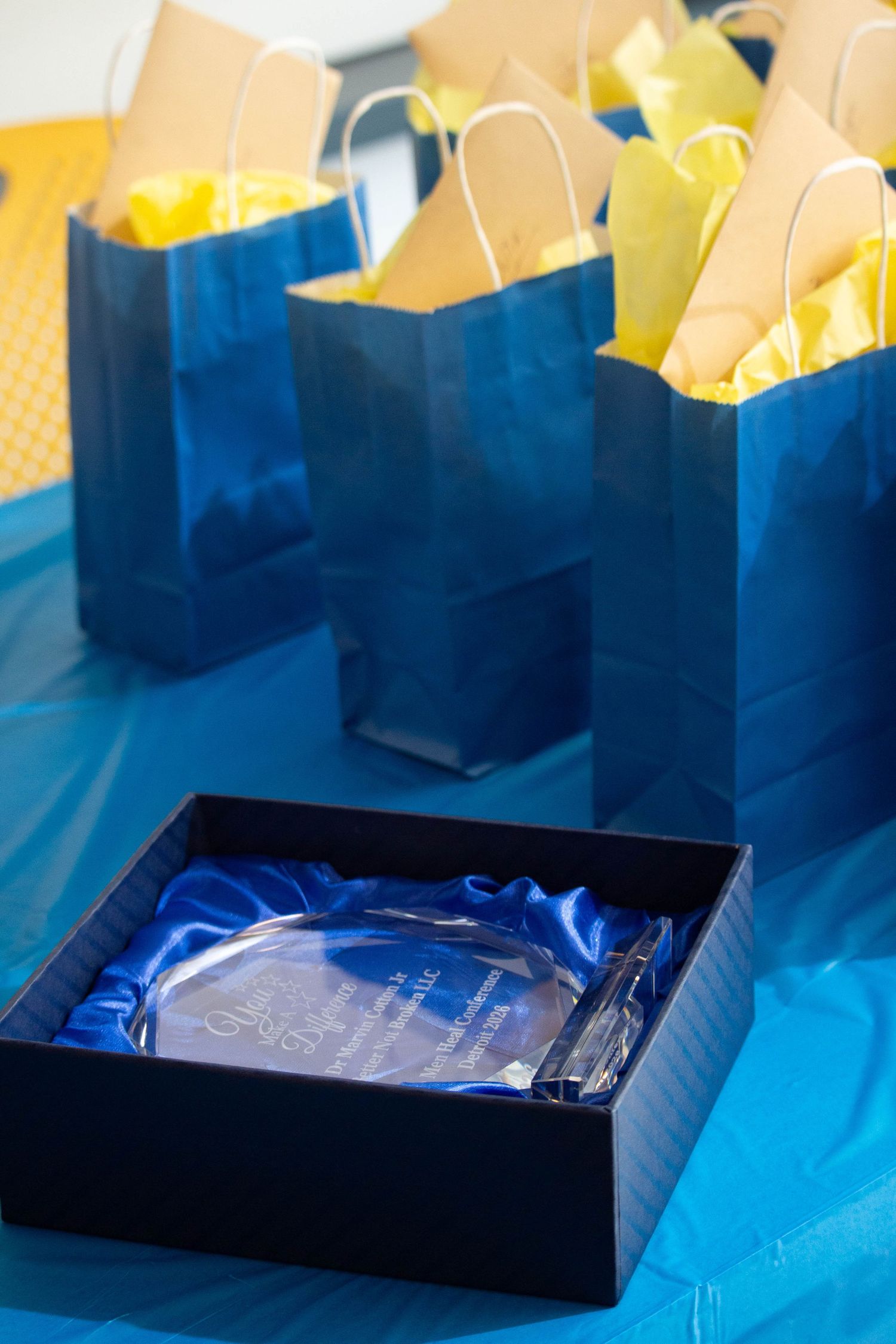Blue gift bags with yellow tissue paper surround a crystal award in a blue satin-lined box on a blue tablecloth.