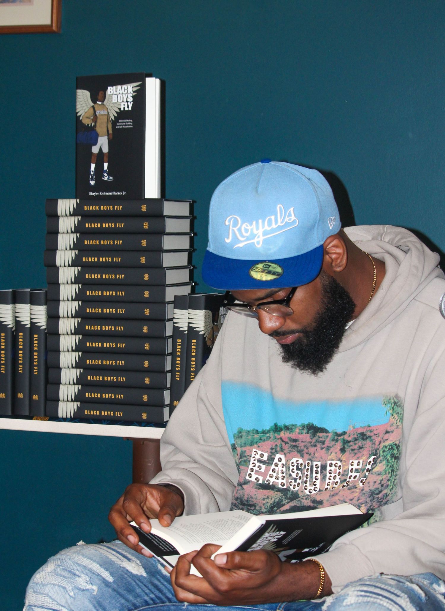 Man in Royals cap reads book near stacked copies of Black Boys Fly at a signing event.