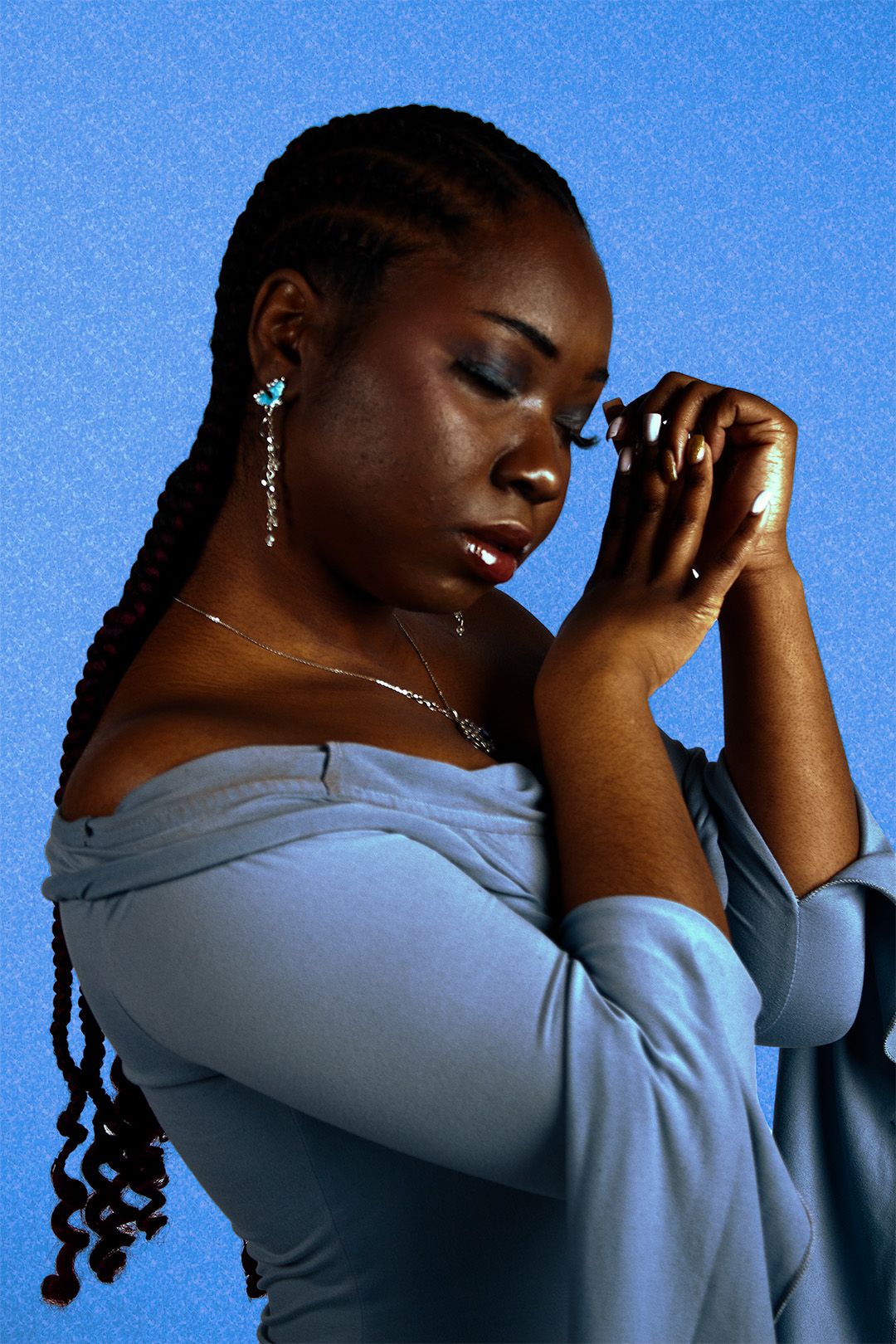 A young Black woman in a blue off-shoulder top poses thoughtfully with hands clasped, wearing dangling earrings against a blue background.