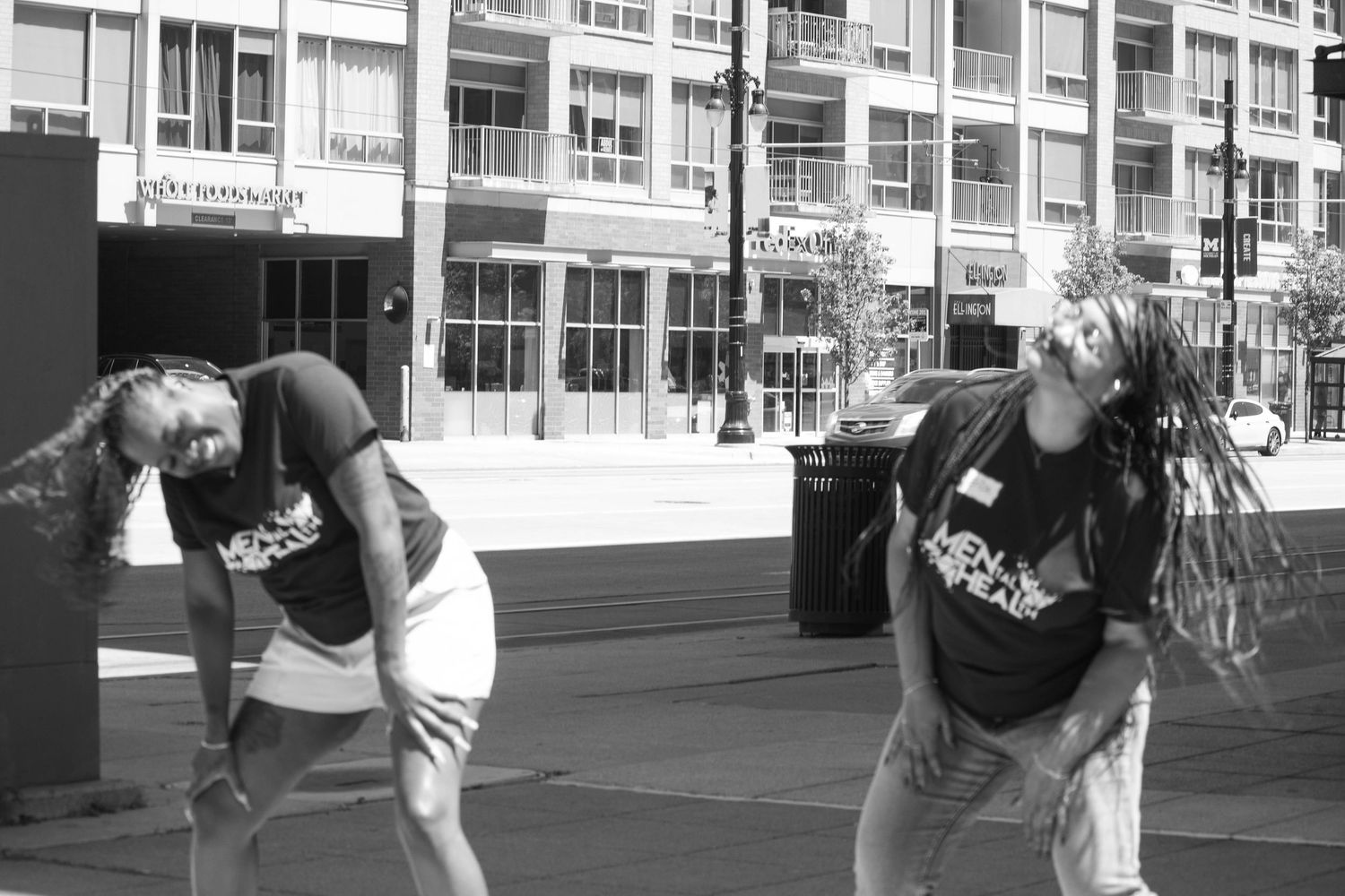 Two people in matching shirts lean back outdoors near a modern building in this black and white street photo.