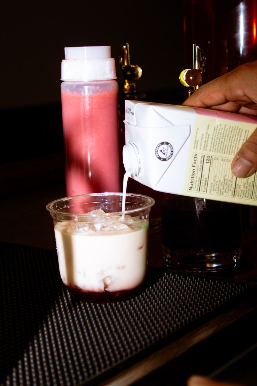 Barista pouring milk from a carton into an iced matcha drink at a cafe counter.