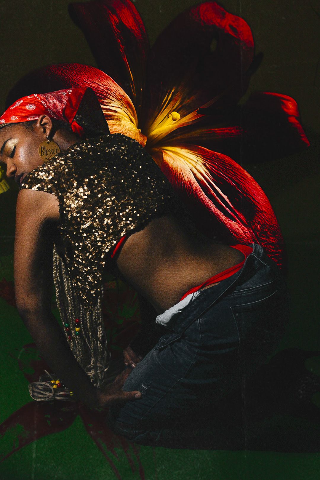 A woman in a gold sequin top poses gracefully with large red tropical flowers against a dark green background.