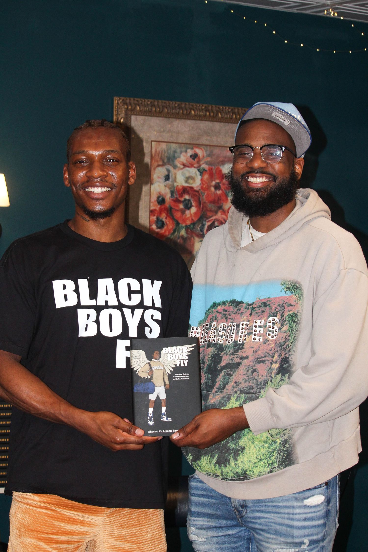 Two smiling men holding a book titled 'Black Boys Fly' in a warmly decorated indoor setting.