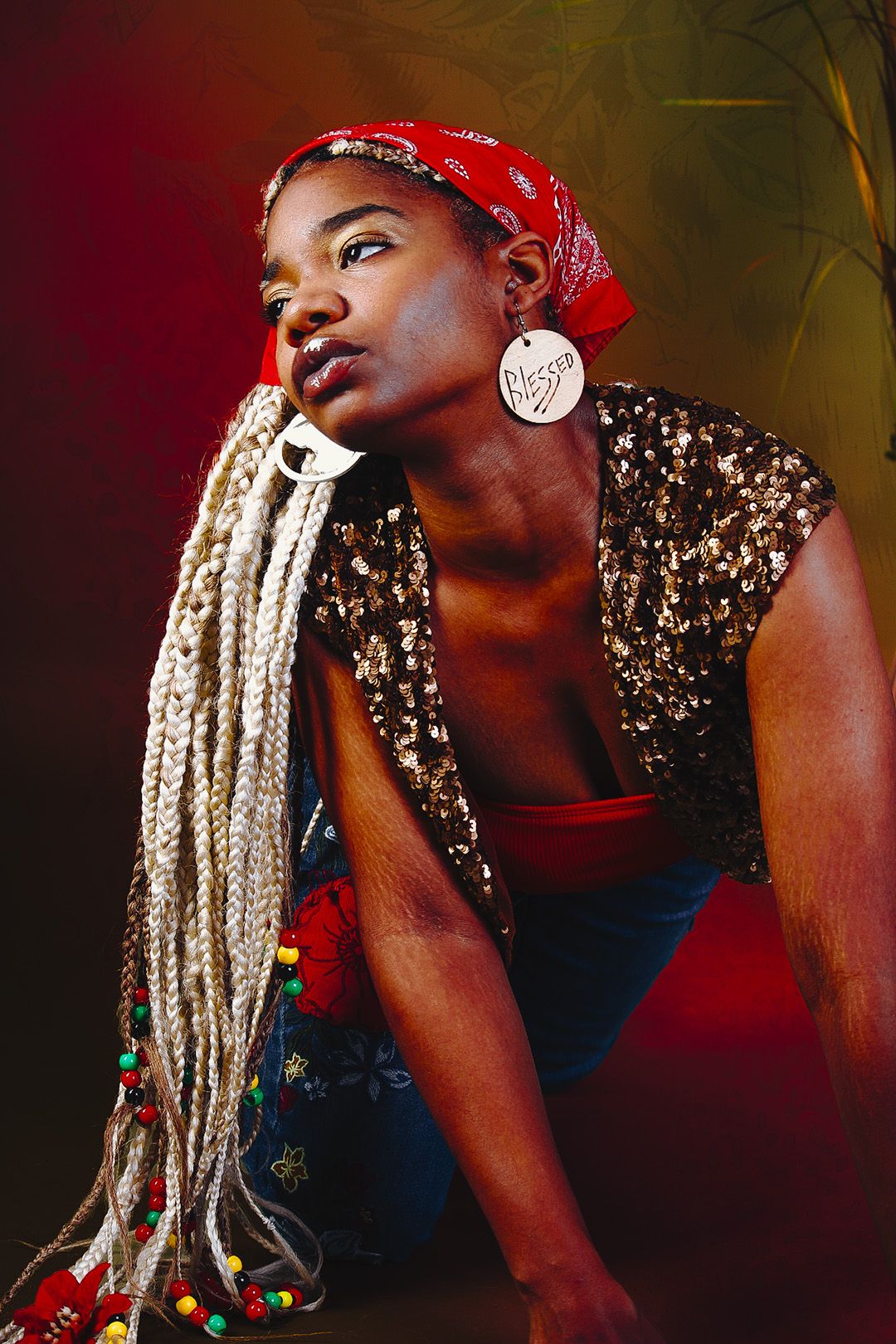 Woman with blonde braids and red bandana wearing sequin top poses confidently in studio.