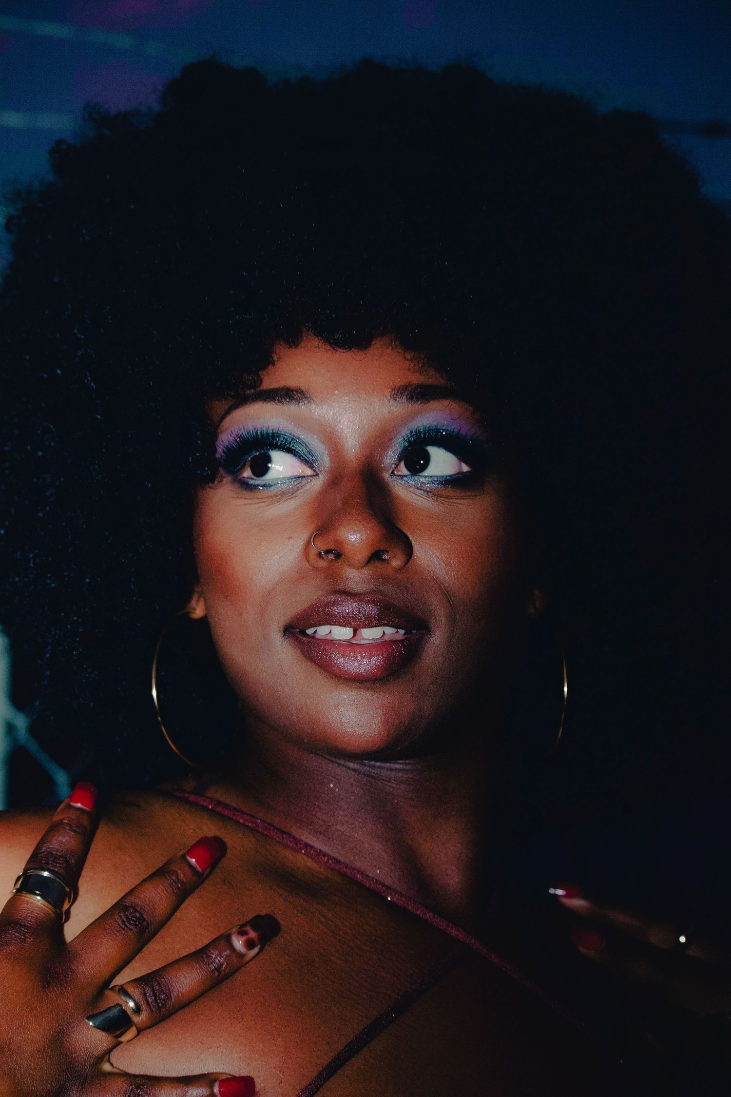 Black woman with afro hair, bold blue eyeshadow, hoop earrings, and rings poses against dark background.