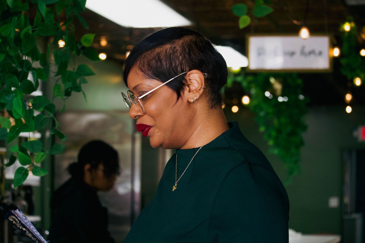 Elegant woman in green top with round glasses and gold necklace at an indoor event with green decor.