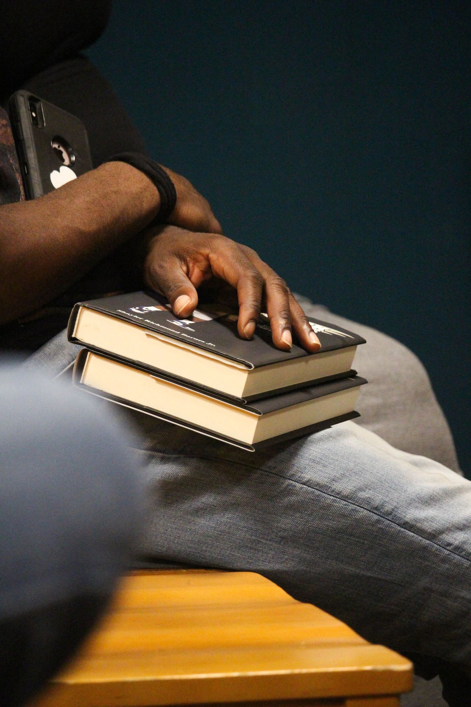 Person sitting with hand resting on two stacked hardcover books on their lap against a teal wall.