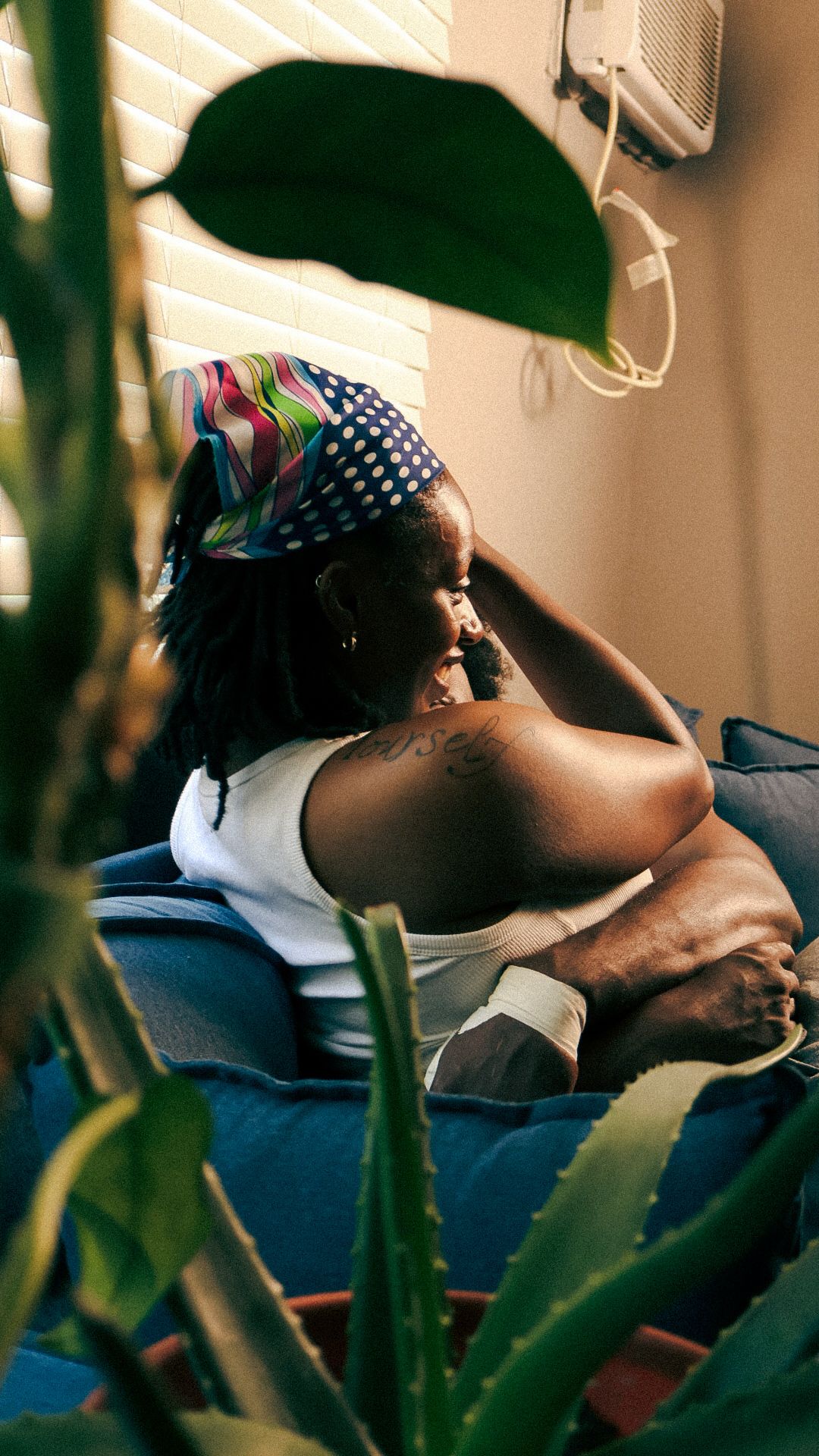 A Black woman with colorful headband relaxes on a blue sofa surrounded by lush green houseplants.