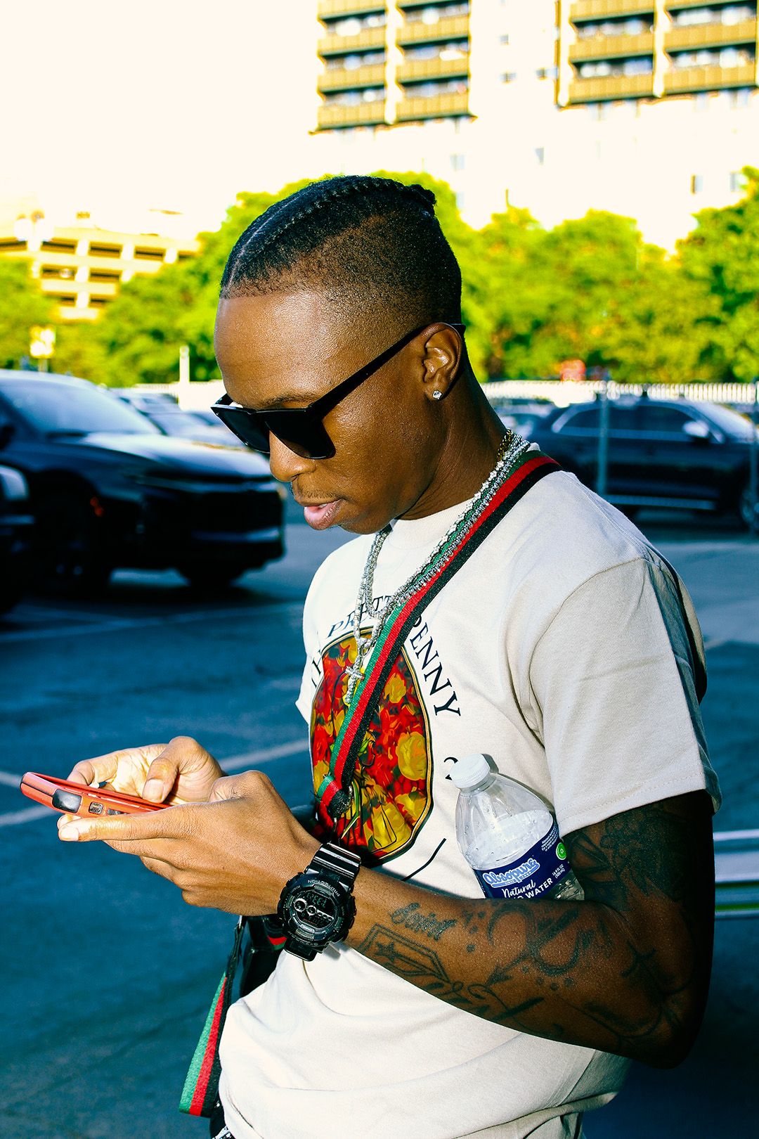 Young man with braids and sunglasses checking his phone outdoors, wearing a white graphic tee and Gucci bag.