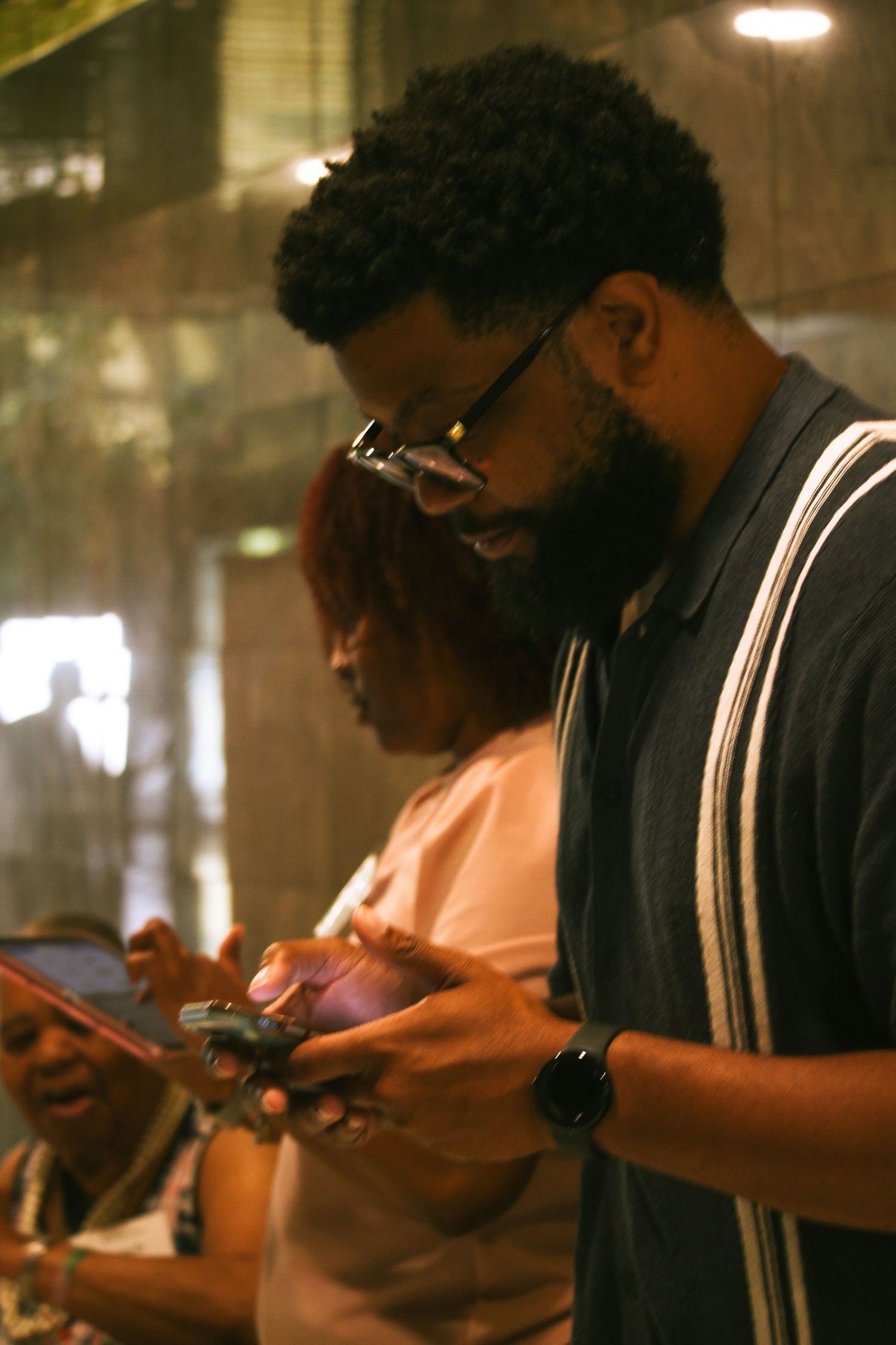 Bearded Black man with glasses looking down at his phone at an indoor event.