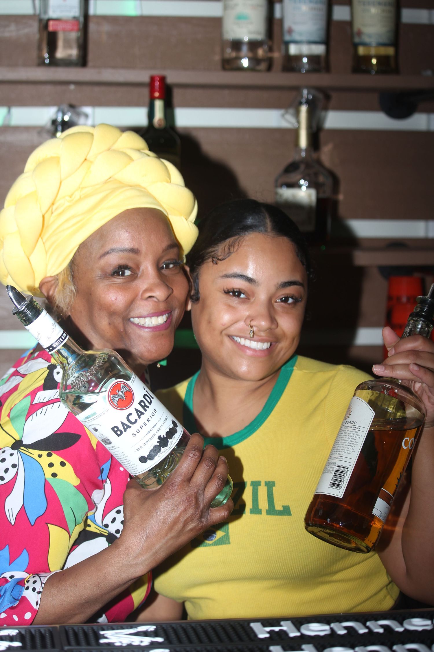 Two smiling women posing with Bacardi and Hennessy bottles behind a bar at a party.