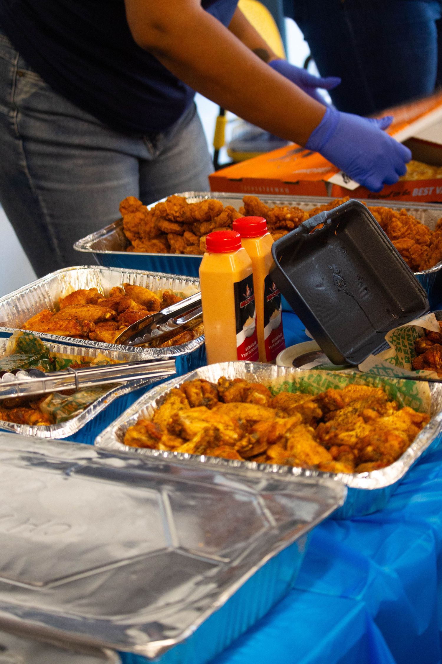 Buffet table with aluminum trays of fried chicken and wings, condiments, served by person wearing blue gloves.