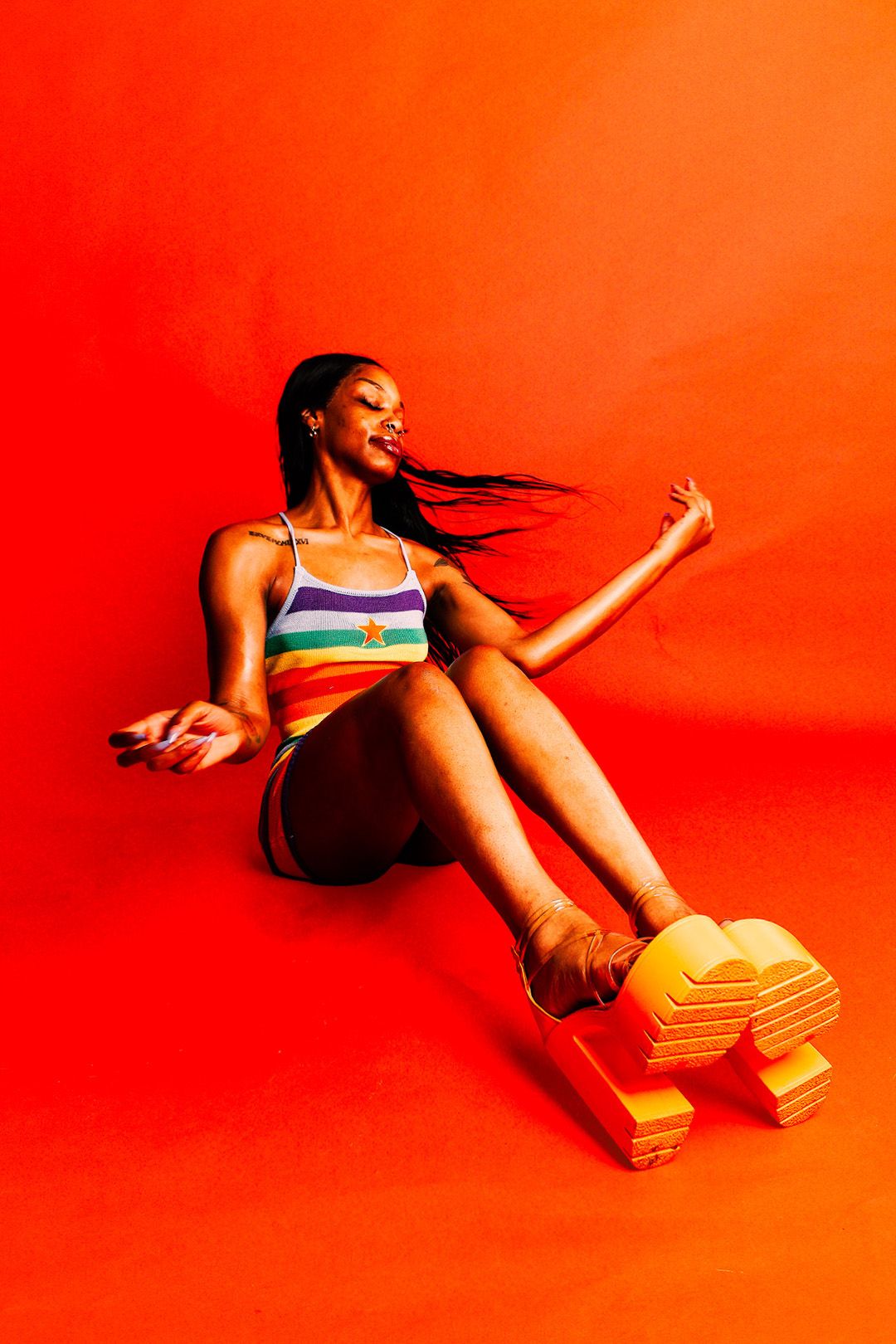 A woman in a rainbow striped top sits on a red background wearing bold yellow platform shoes.
