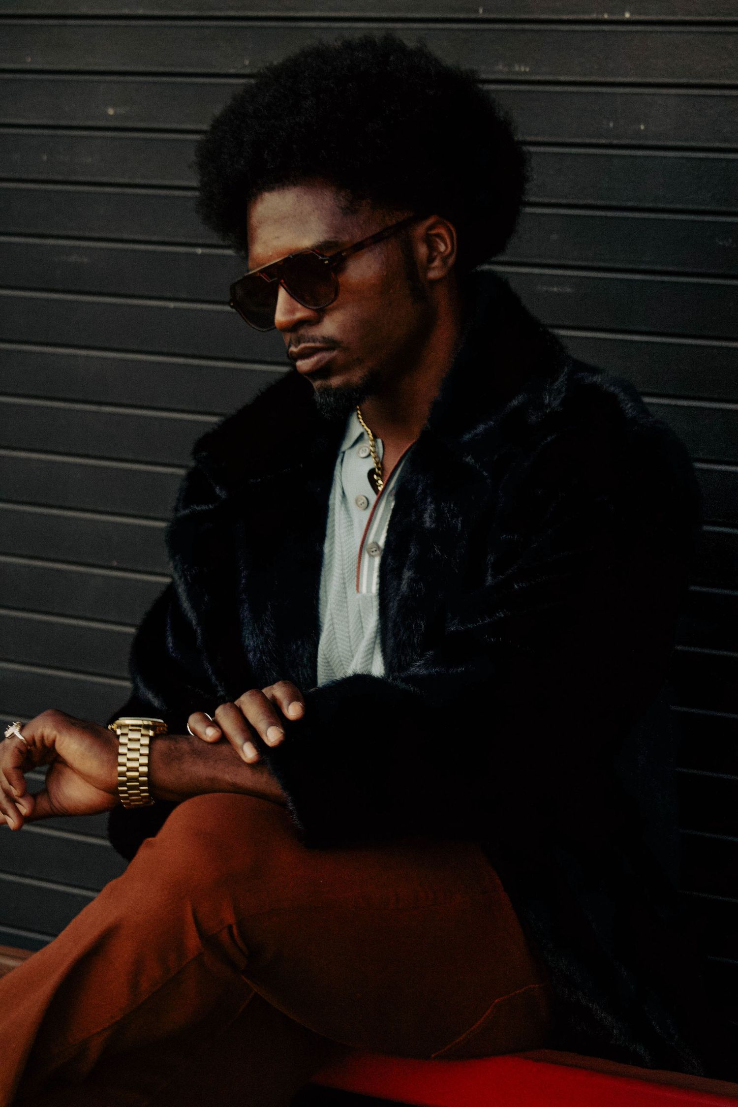 Stylish young Black man with afro wearing sunglasses, fur coat, and gold watch, sitting against a dark wall.