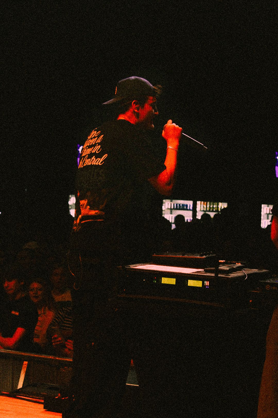 Male performer in black graphic tee singing into microphone on dark concert stage.