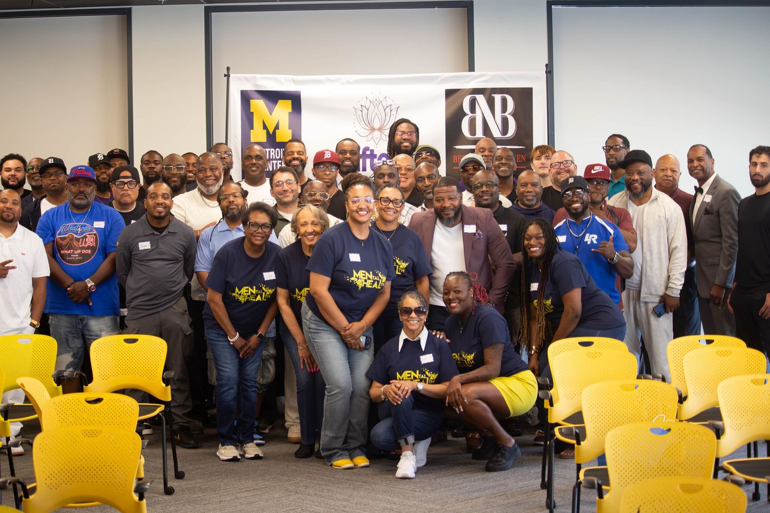 Large group of diverse men posing together indoors at a community event with yellow chairs and organization banners.