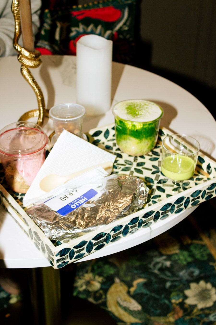 A decorative mosaic tray holds matcha drinks, candles, and snacks on a white table with floral decor.