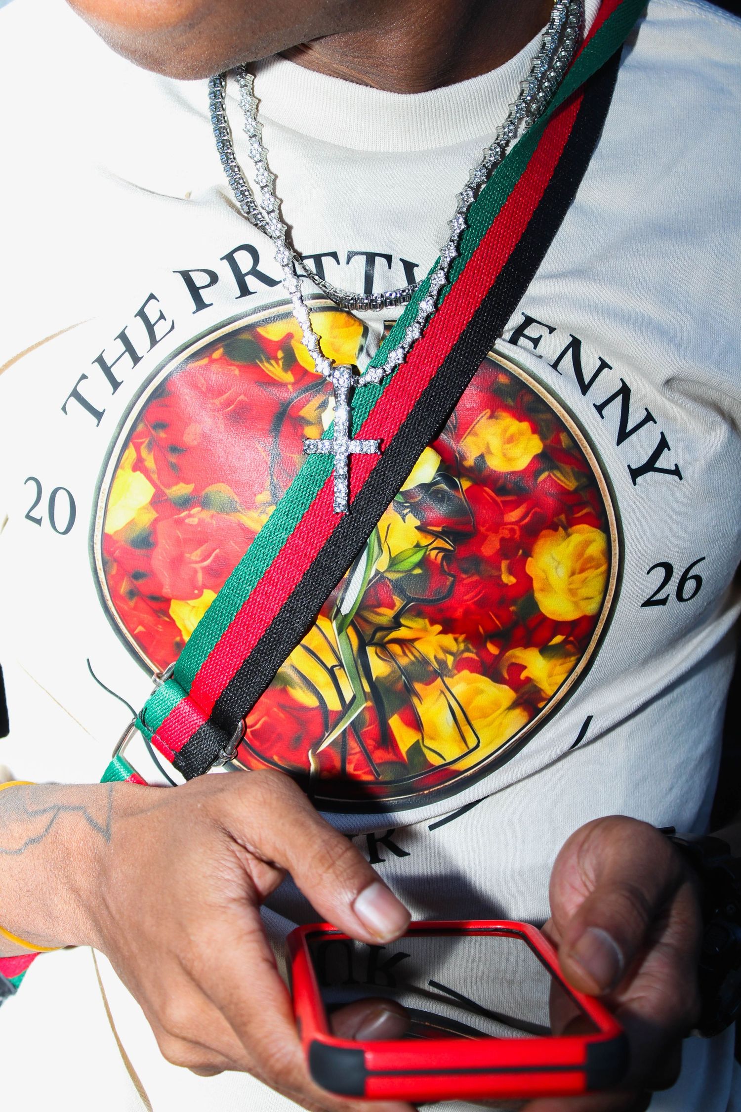 Man wearing Gucci crossbody bag and cross necklace over floral graphic tee, holding red phone.