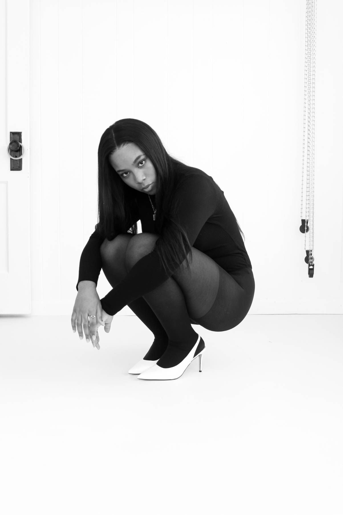 Black and white photo of a woman in all-black outfit crouching gracefully in white heels against a white background.