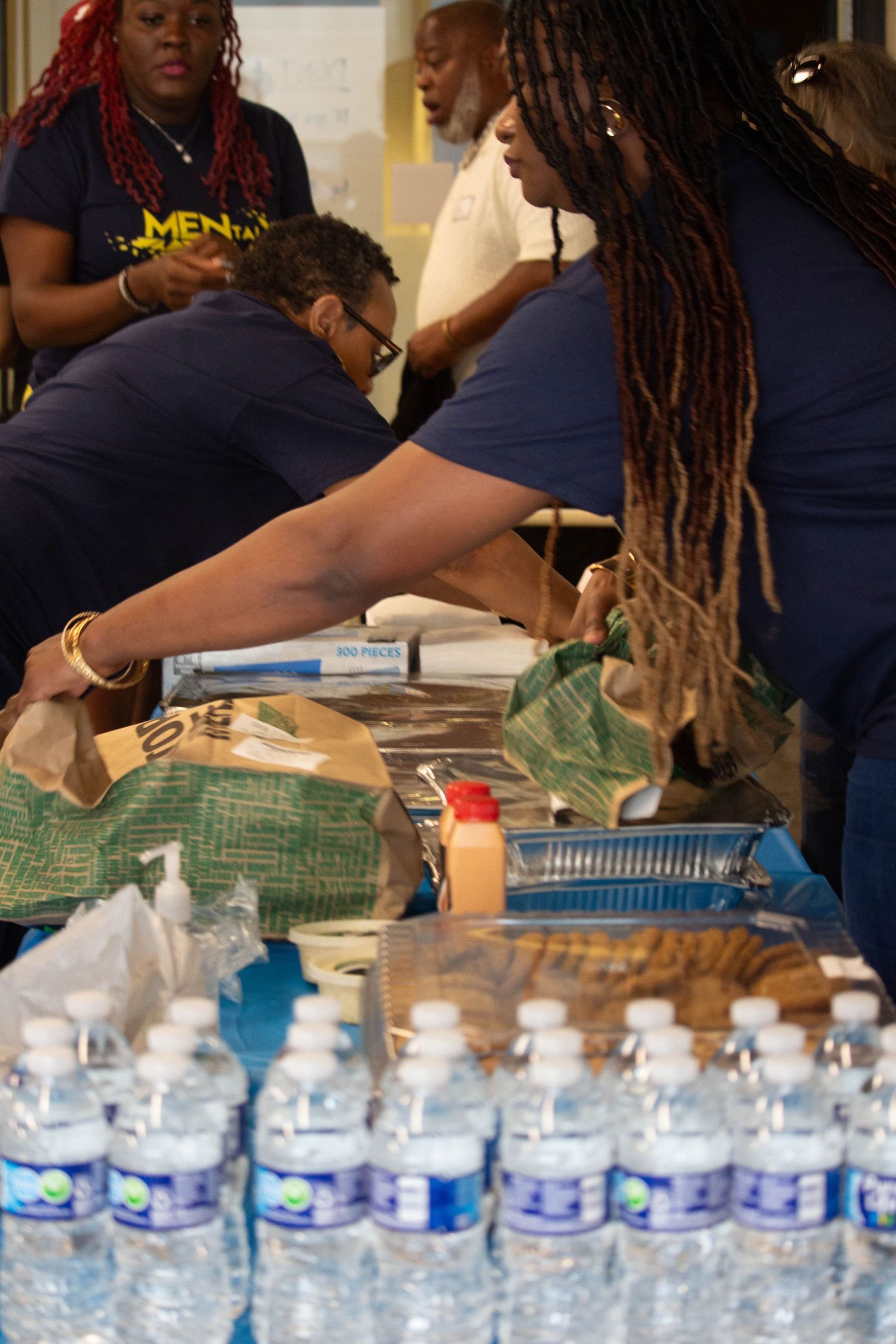 Volunteers in navy blue shirts serve food and water bottles at a community event or food drive.