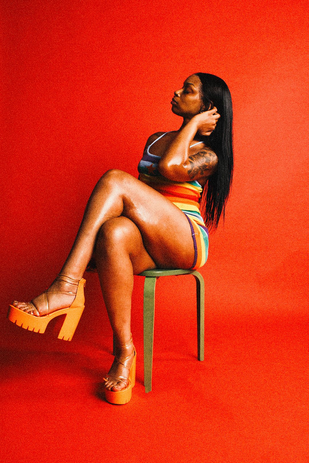 A Black woman in a colorful striped bodysuit and orange platform heels sits on a stool against a bold red background.