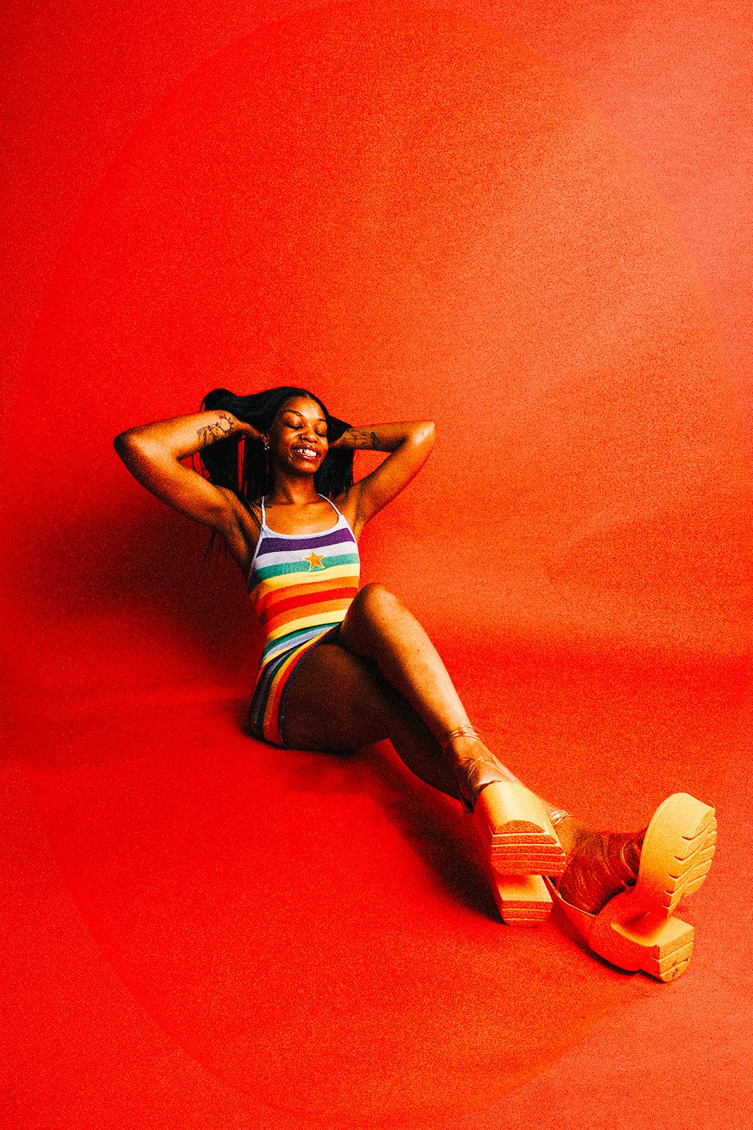 Smiling Black woman in rainbow striped bodysuit and platform shoes relaxes on floor against red background.
