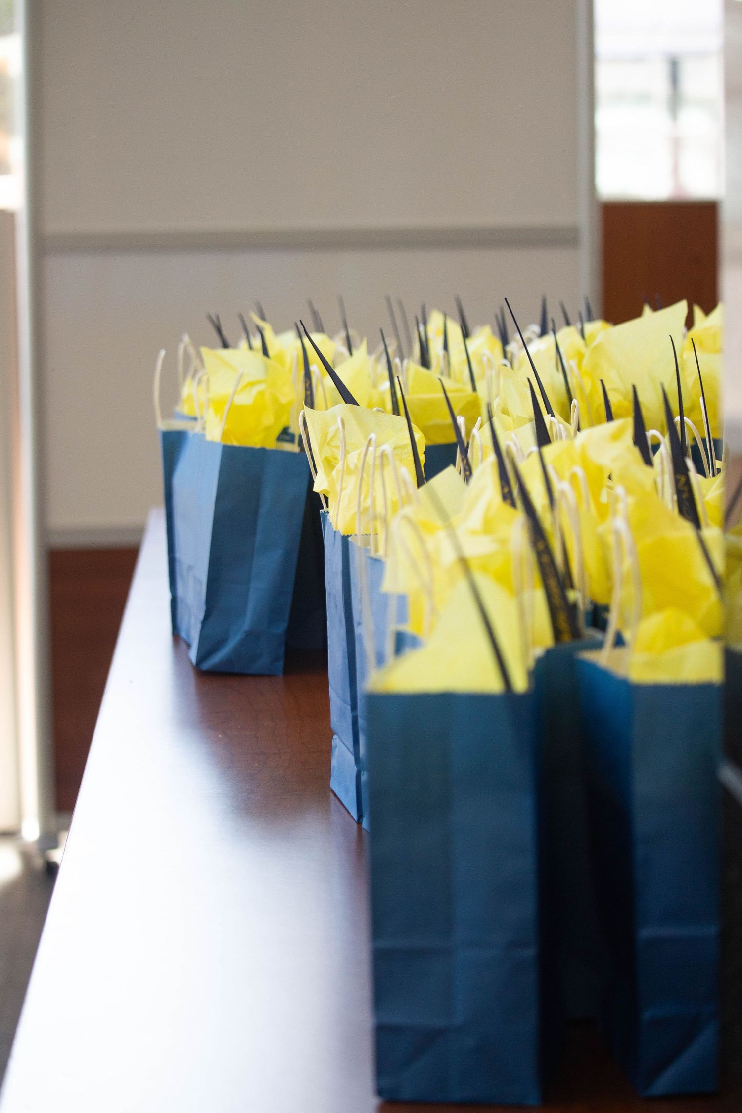 Blue gift bags filled with yellow tissue paper and black handles arranged in rows on a wooden table.