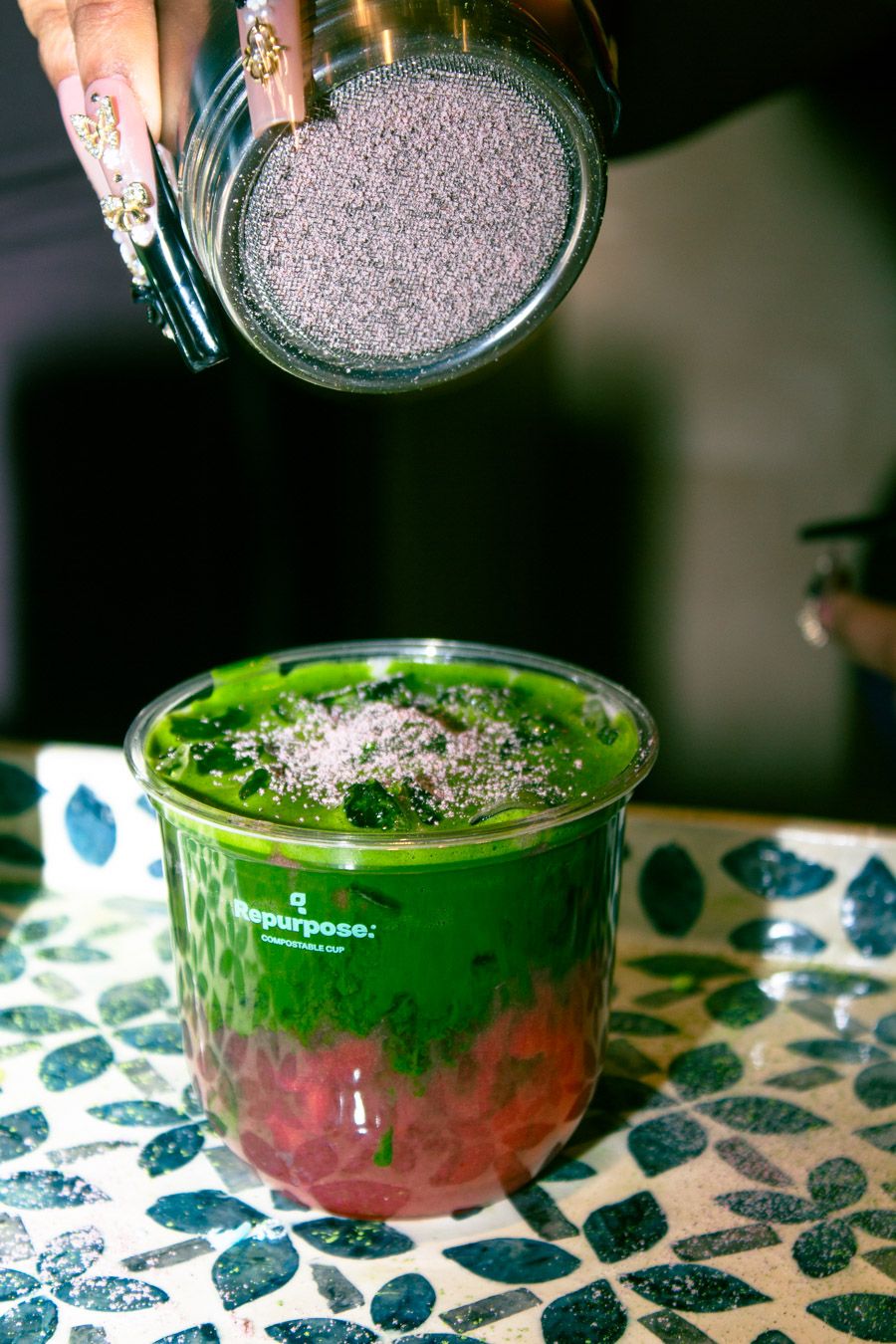 Vibrant green matcha drink being topped with purple powder in a clear plastic cup on a mosaic table.