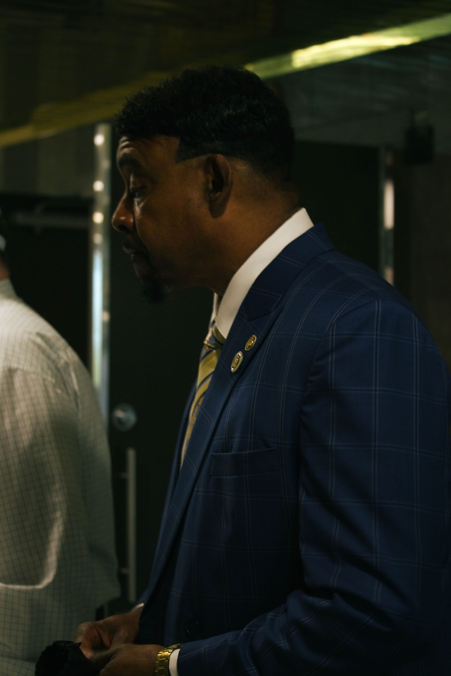 Well-dressed Black man in a blue plaid suit and gold tie seen in profile at an indoor event.