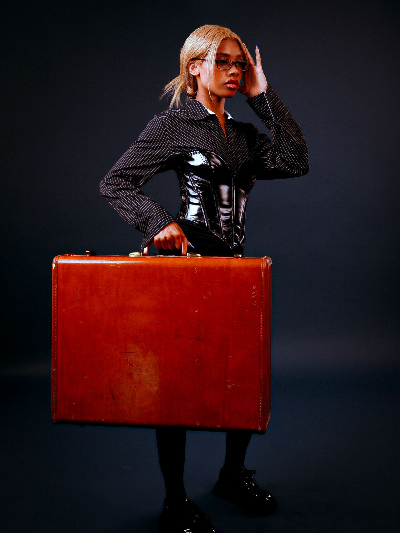 Woman in pinstripe blazer and corset holding a large vintage brown suitcase against dark background.