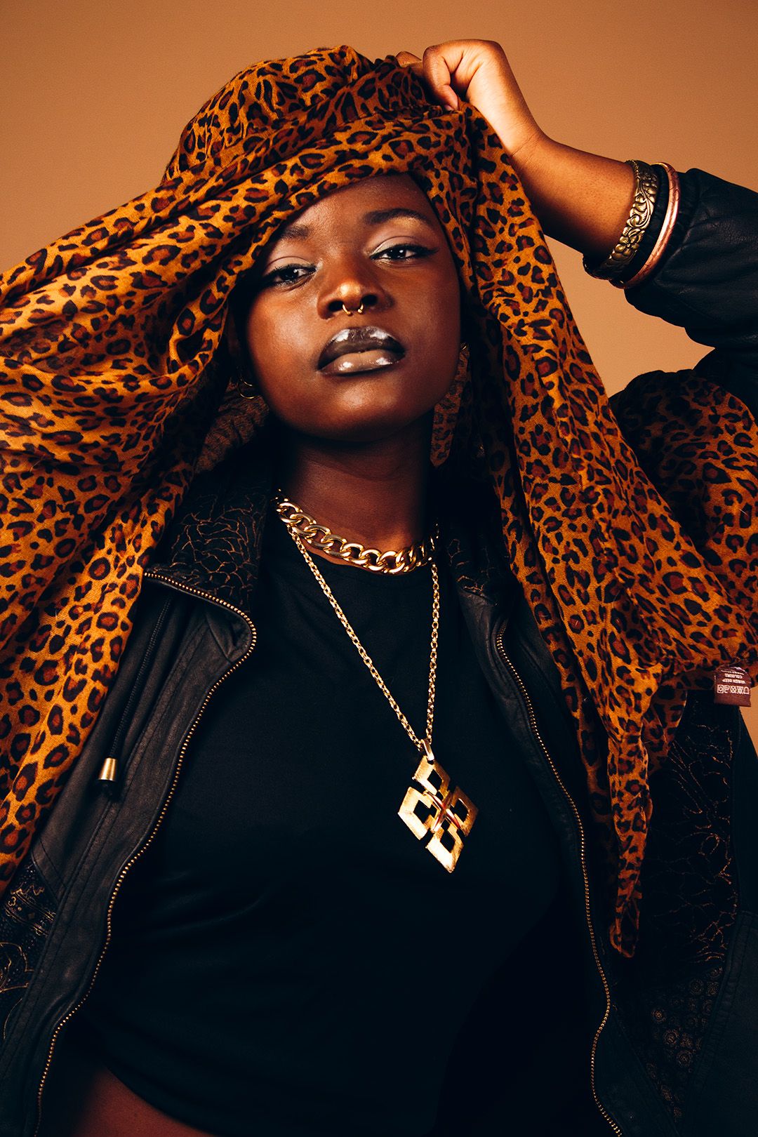 Black woman in leopard print headwrap and gold chain necklace poses against warm brown background.