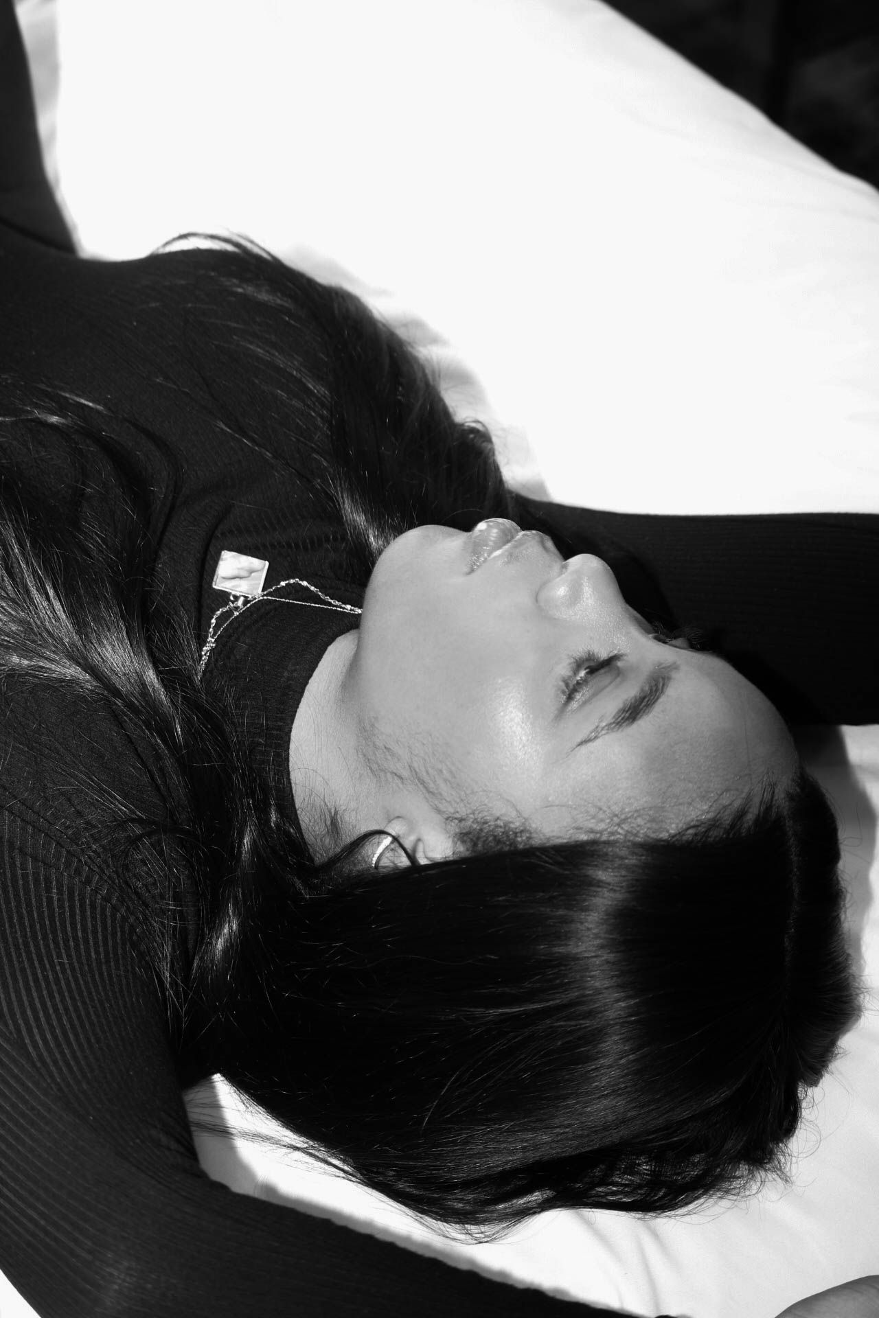 Black and white photo of a woman with long dark hair lying down, eyes closed, wearing earphones and a dark top.