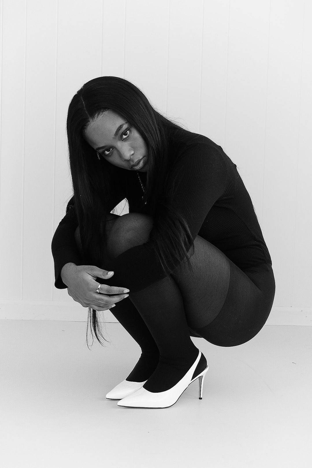 Black and white photo of a woman in black outfit and white heels crouching gracefully against a white background.