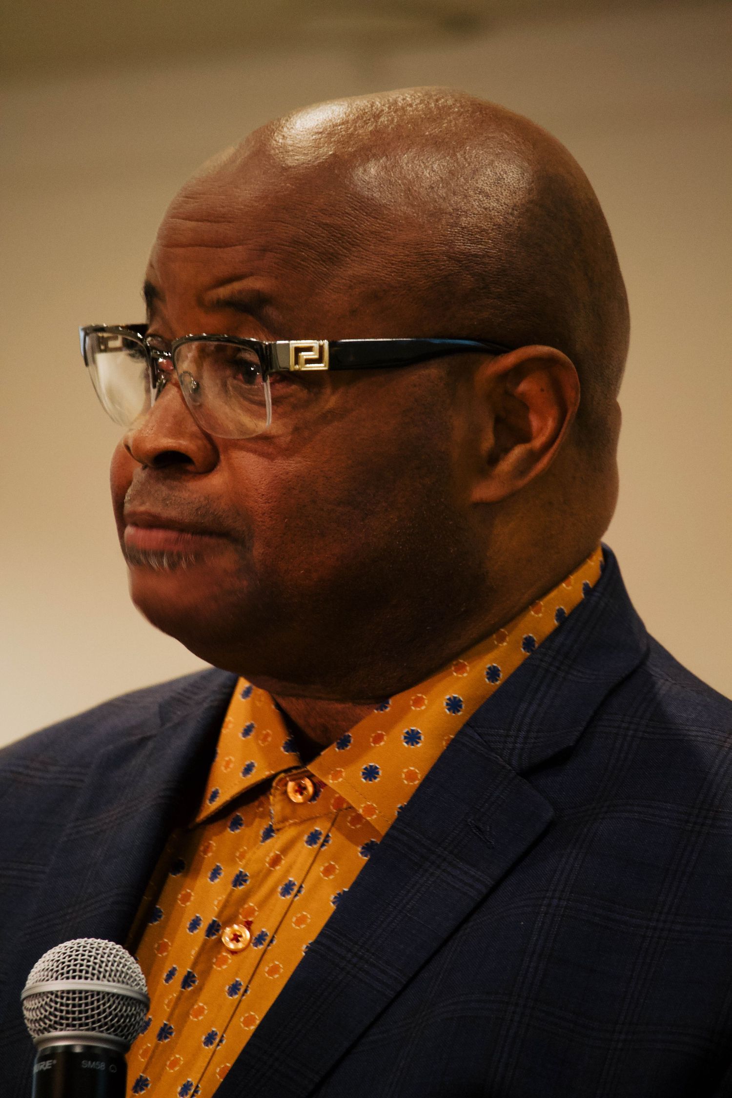 Bald Black man wearing Fendi glasses, a gold polka dot shirt, and blue blazer speaking at an event.