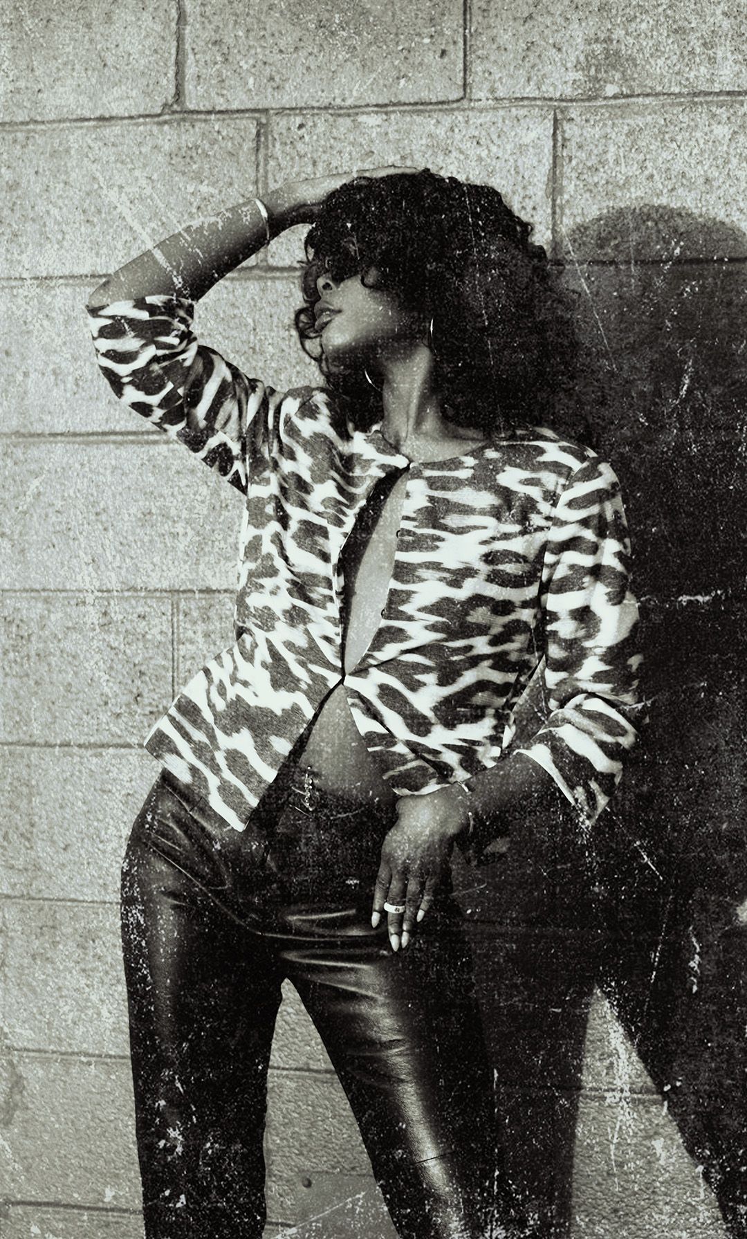 Black and white photo of a woman in leopard print jacket and leather pants posing against a brick wall.