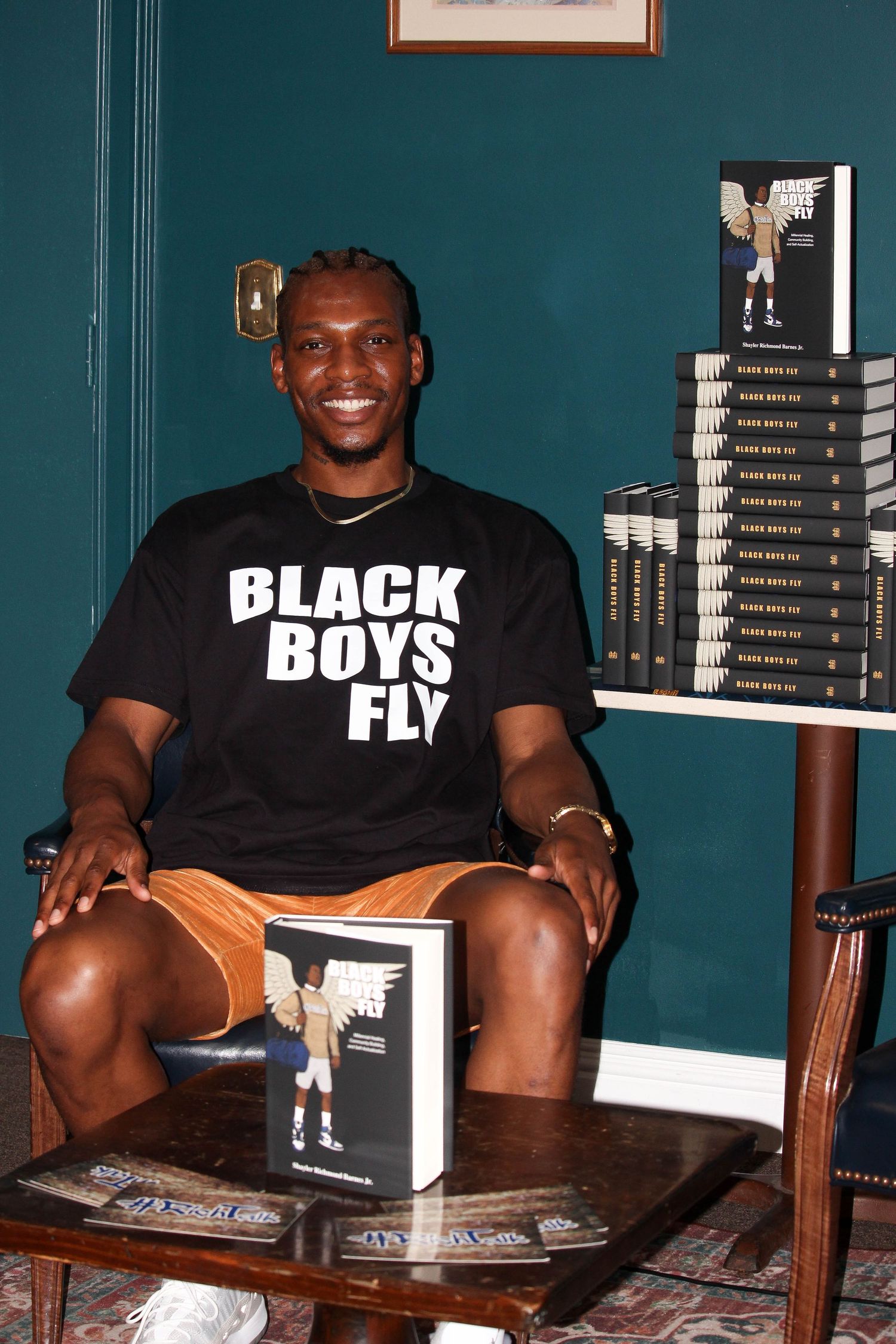 Man in Black Boys Fly t-shirt sits smiling beside stacked copies of his book at a signing event.