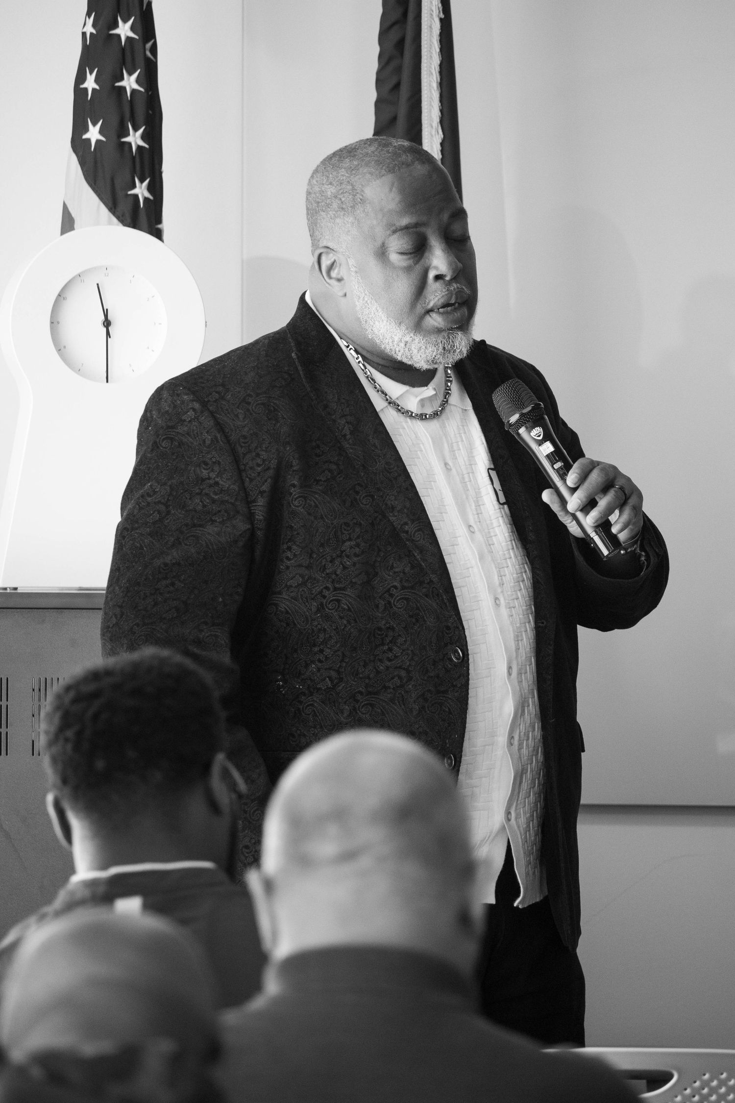 Black and white photo of an older man speaking into a microphone at an indoor event.