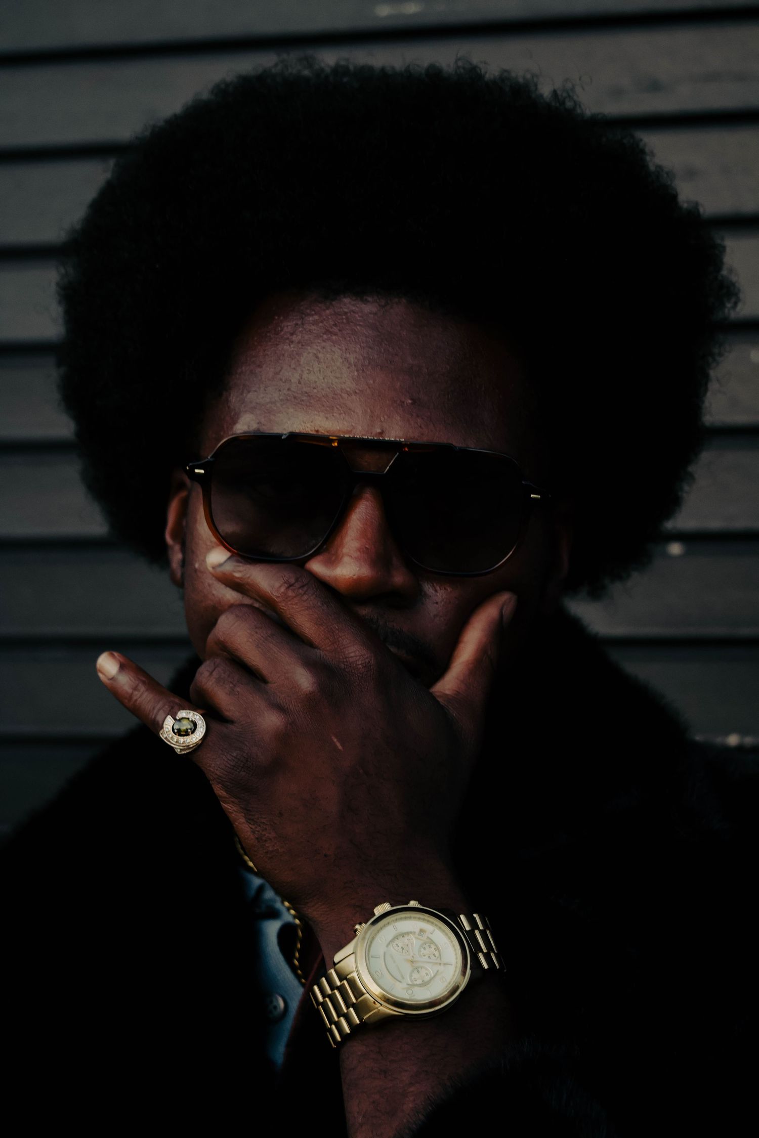 A stylish Black man with an afro wearing sunglasses, a gold watch, and ring, smoking a cigarette in dark lighting.