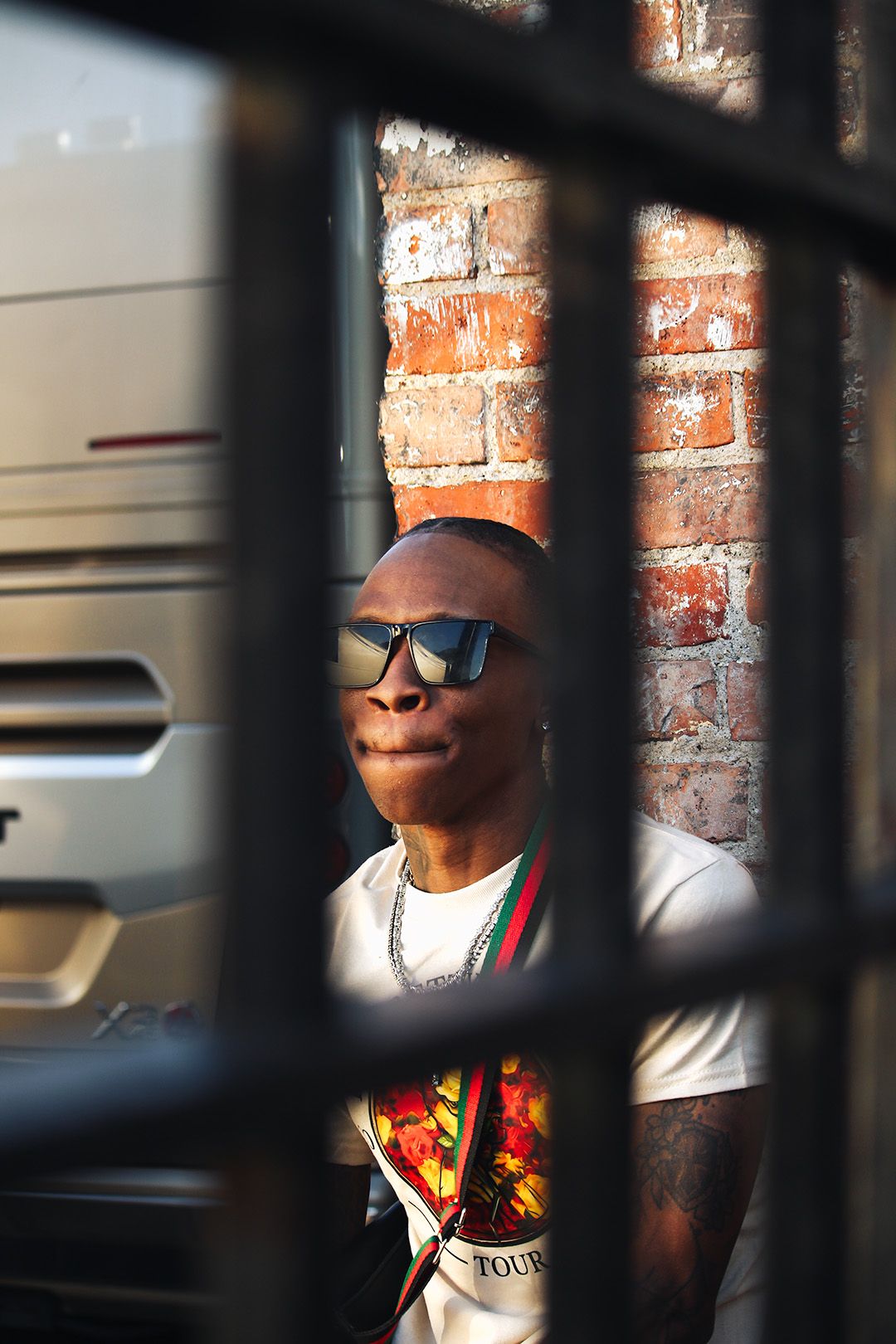 Man wearing sunglasses and white graphic tee stands against brick wall, viewed through metal bars in urban setting.