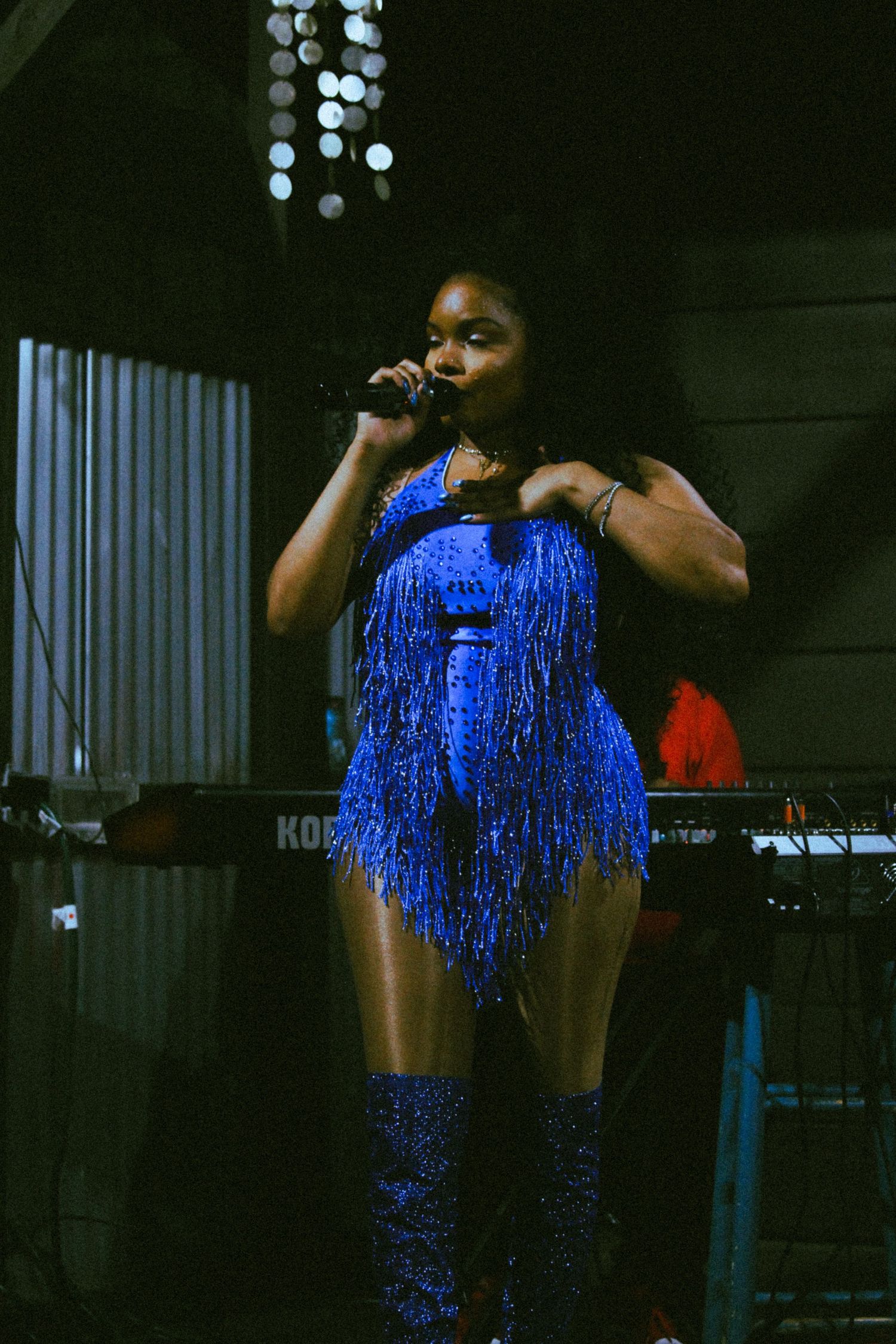 Woman performing on stage in a blue fringed sequin bodysuit and boots, singing into a microphone under stage lights.