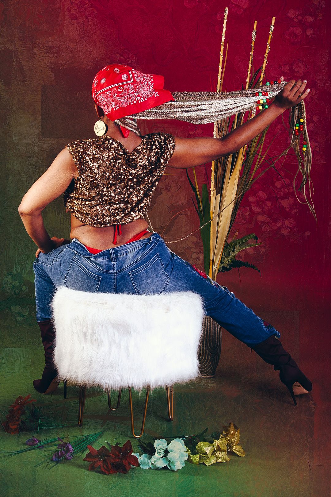 Woman in red bandana, sequin top, and jeans poses on white fur chair with braids extended, surrounded by flowers.