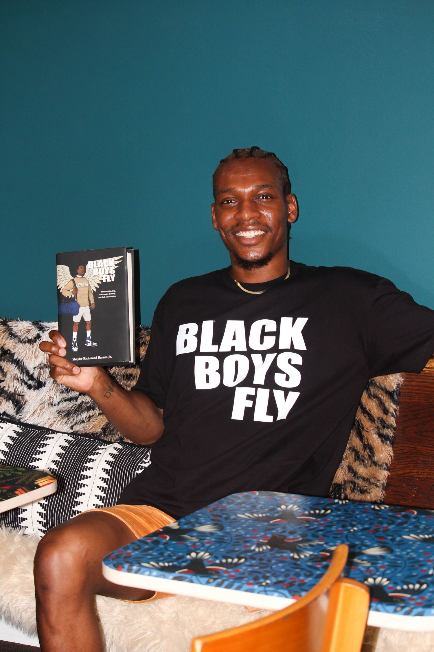 Man smiling holding a book wearing a Black Boys Fly t-shirt seated on a couch.