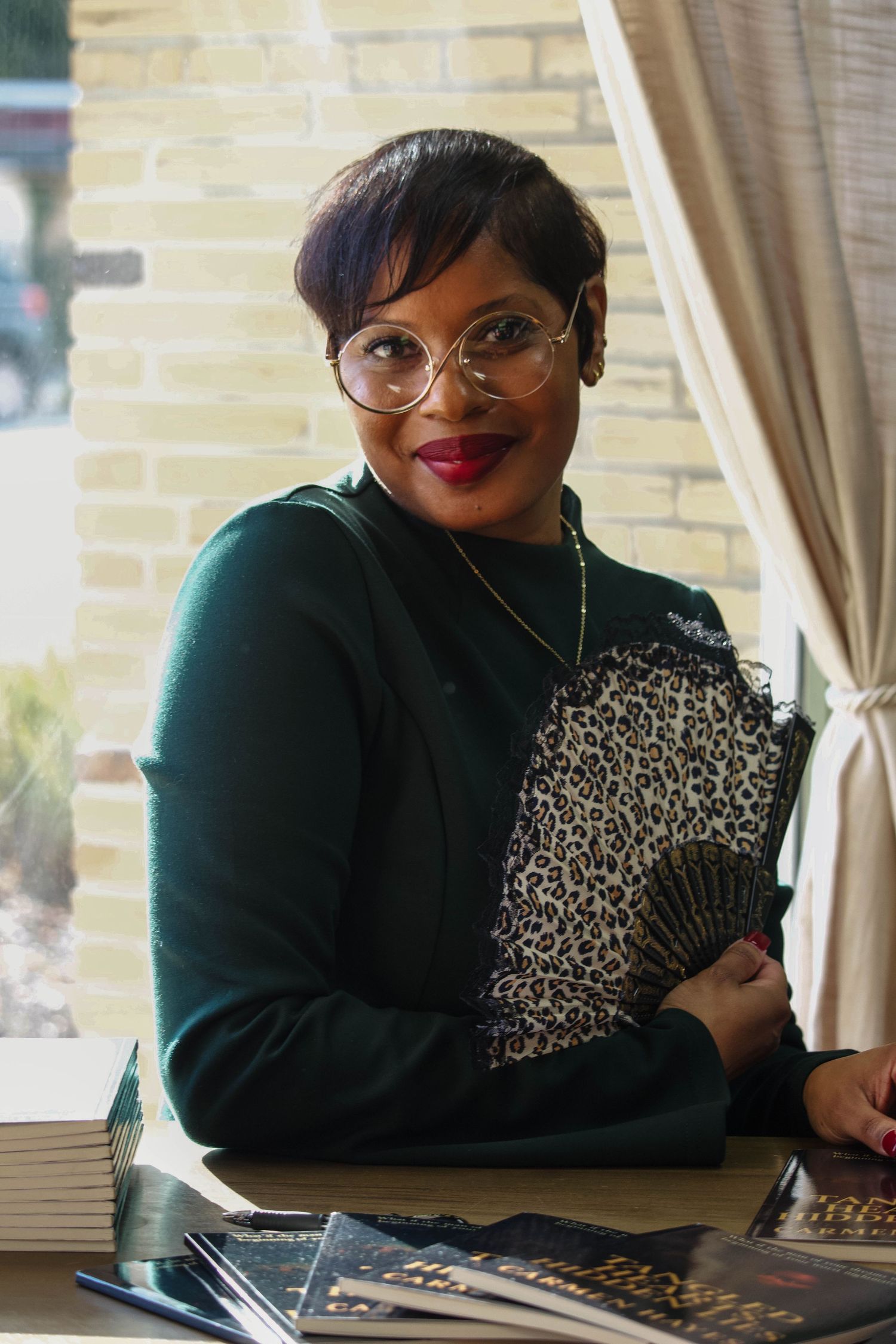 Smiling Black woman author with glasses and red lips holds a fan beside stacked books at a signing event.