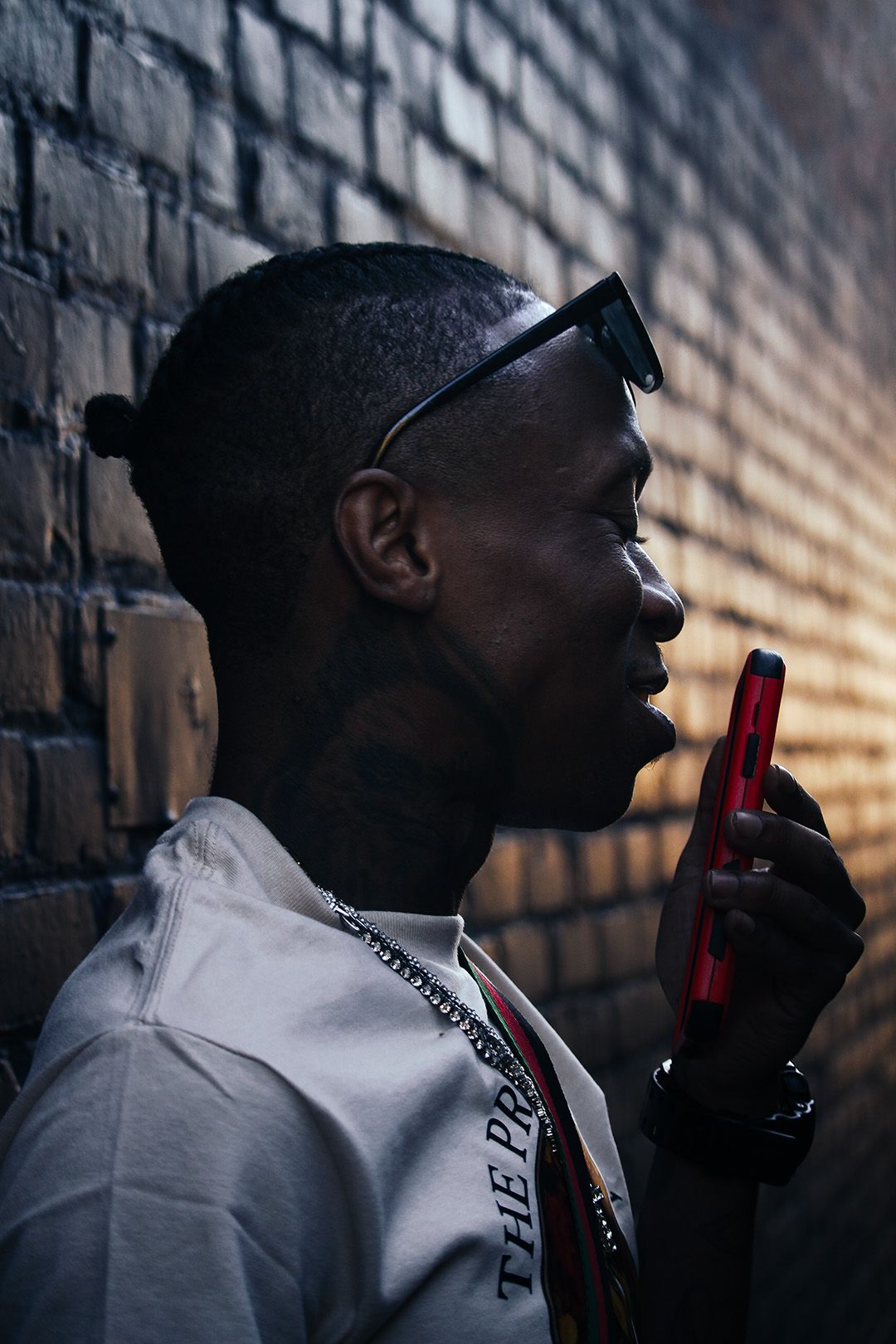 Young man in sunglasses holding red phone against brick wall in dramatic lighting.