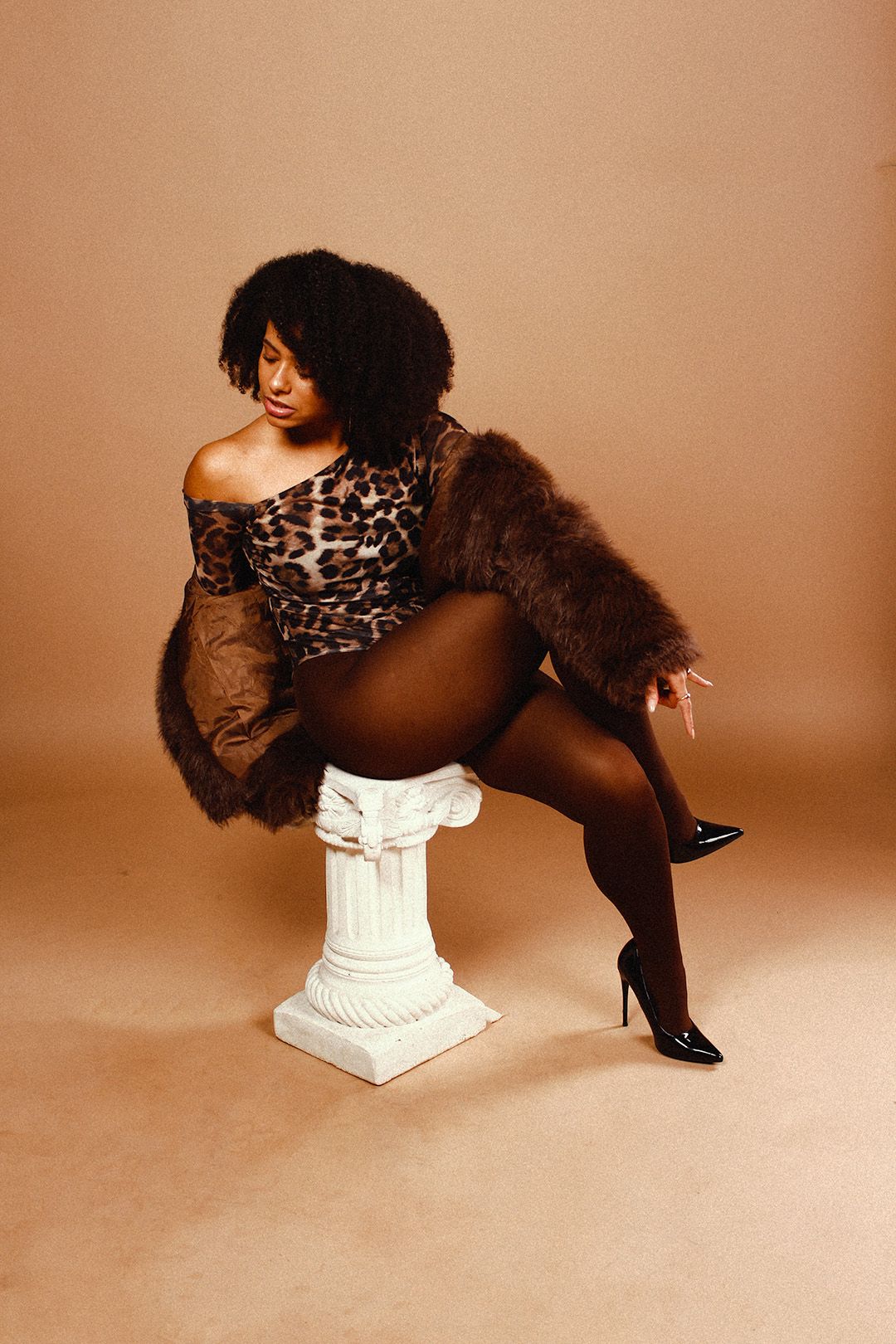 Woman in leopard bodysuit, fur coat, and black heels posing on a white pedestal against a beige background.