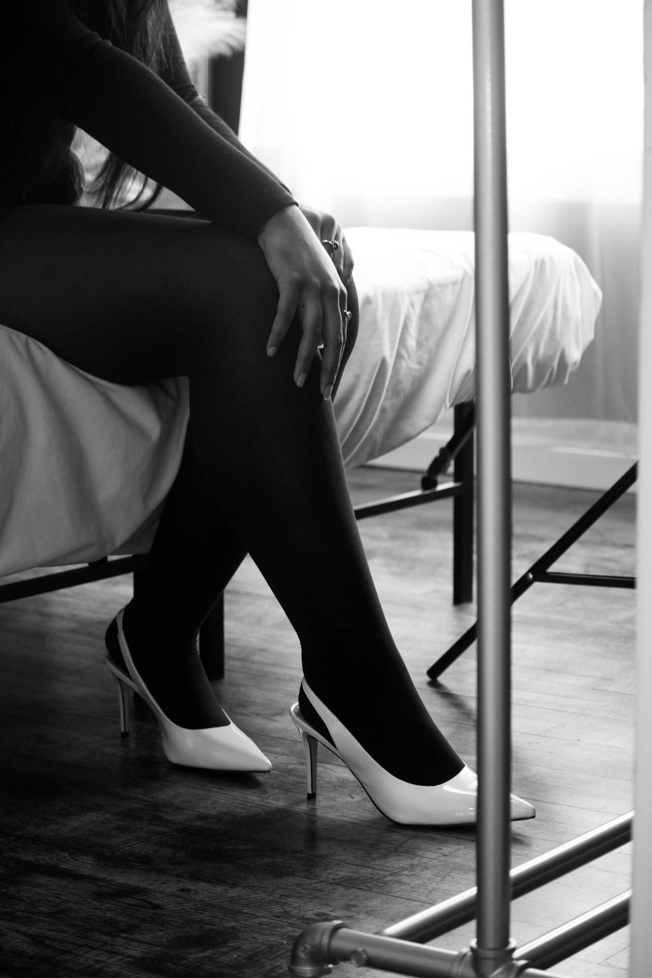 Black and white photo of a woman sitting on a bed wearing high heels and dark stockings.