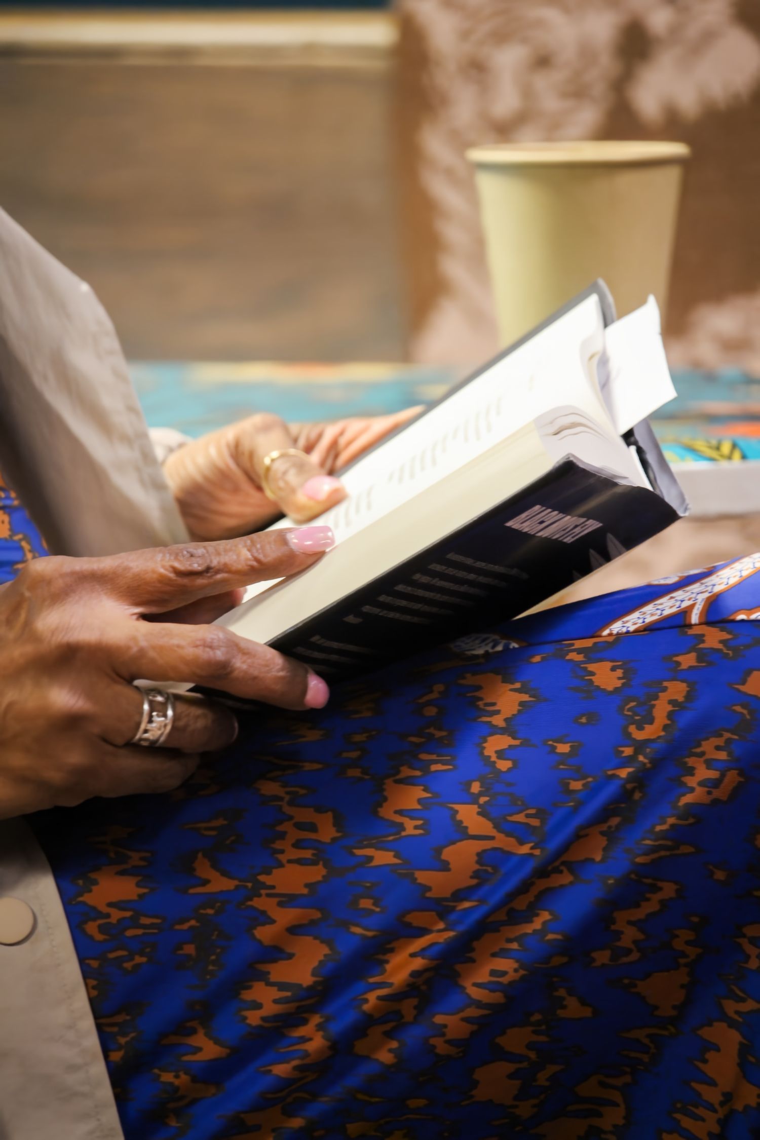 Elderly person reading a book while seated, wearing a blue patterned skirt and a silver ring on their finger.